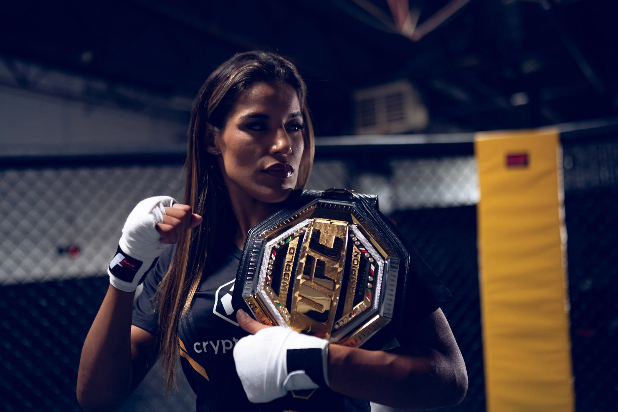 UFC Champion Julianna Pena holding a championship belt in a fighting cage, wearing fight gloves and sporting long hair, with a serious expression.