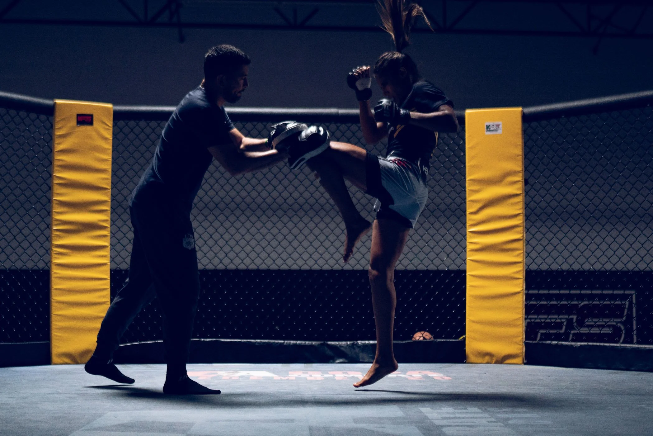 UFC Champion Julianna Pena practicing kickboxing in a UFC-style octagon, with a trainer holding a focus mitt for her to strike, in a dimly lit gym.