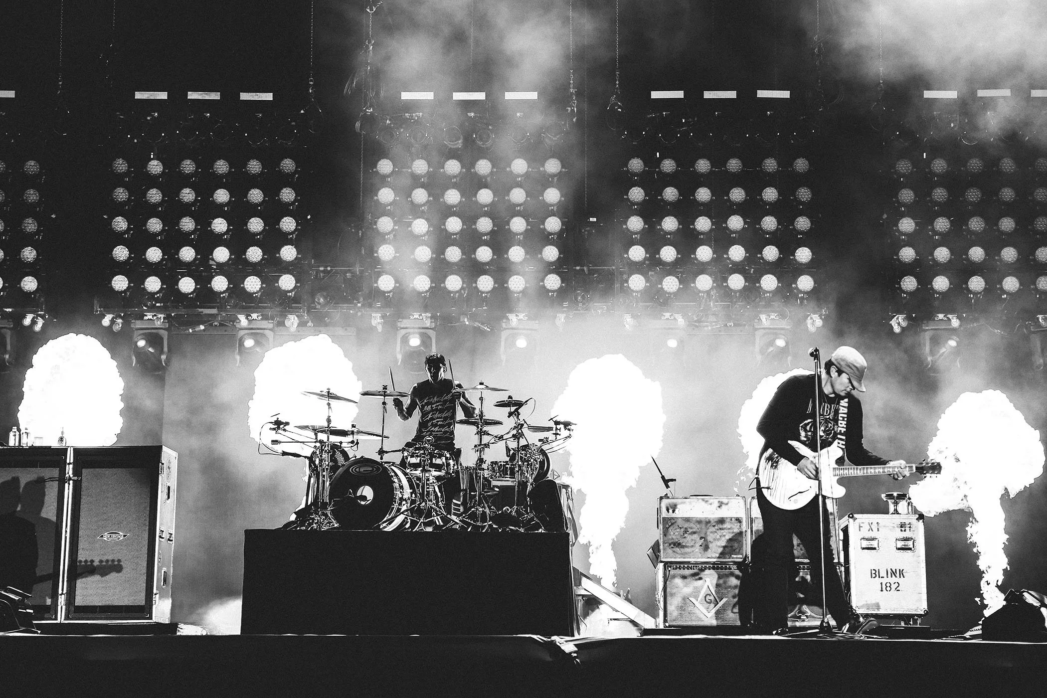 Black and white photo of a Travis Barker performaning with two musicians on stage, one playing drums and the other playing guitar, with smoke and bright stage lights in the background.