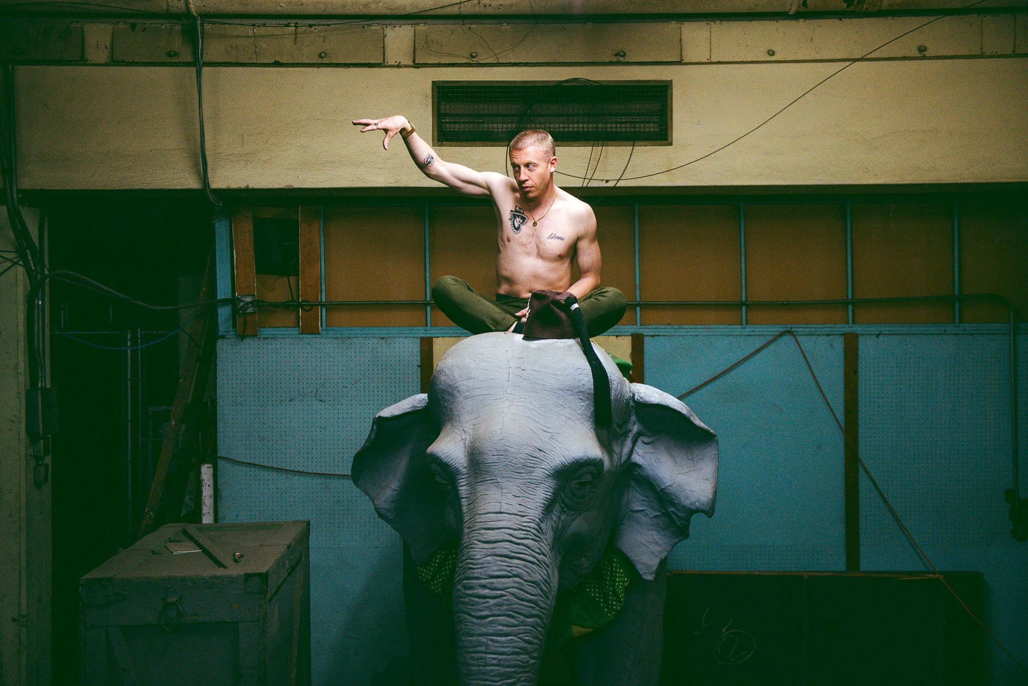 Macklemore sitting cross-legged on a large elephant statue inside a room with exposed wiring and a yellow-green wall.