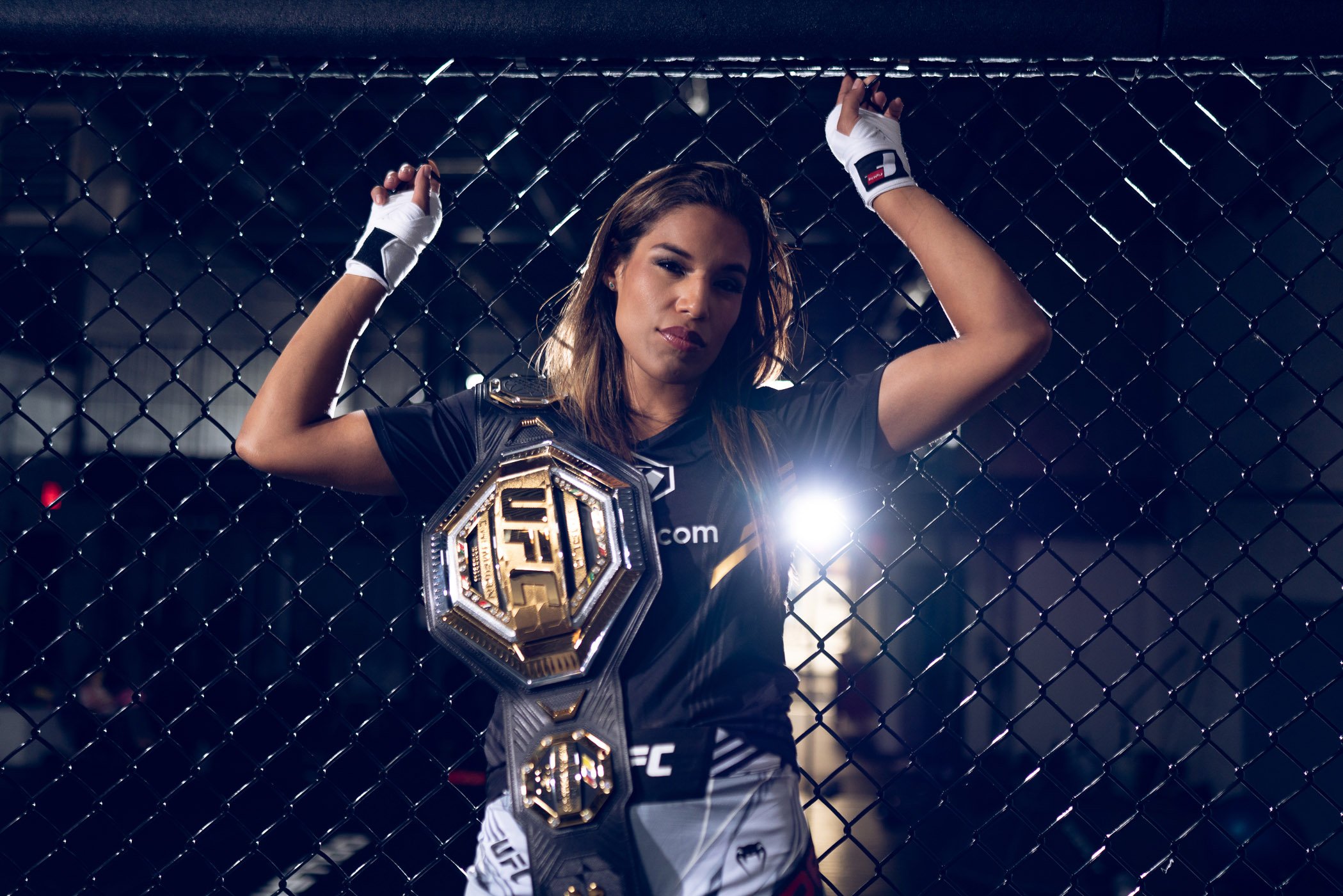UFC Champion Julianna Pena standing inside a cage with her arms raised, wearing a championship belt over her shoulder, black shorts, and fighting gloves, with a serious expression.