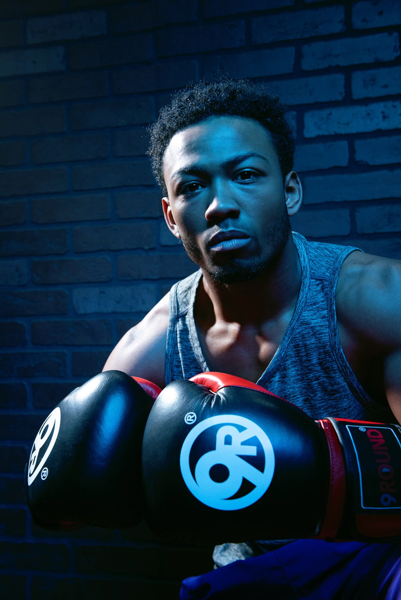 A campaign for 9Round featuring an athlete wearing boxing gloves and a gray tank top, posing confidently in front of a dark brick wall.