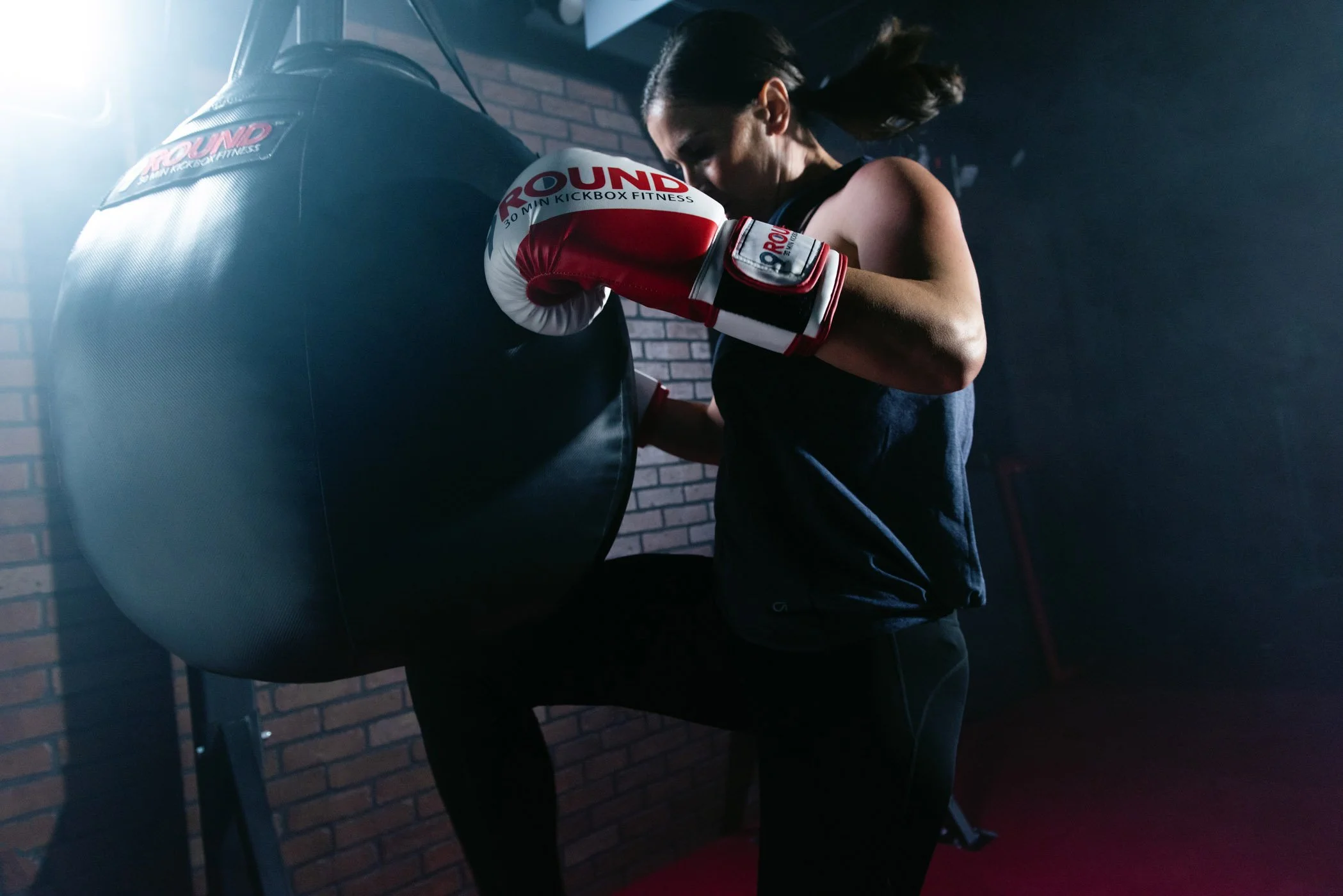 A campaign for 9Round featuring a female athlete wearing boxing gloves training with a punching bag in a gym with brick walls.