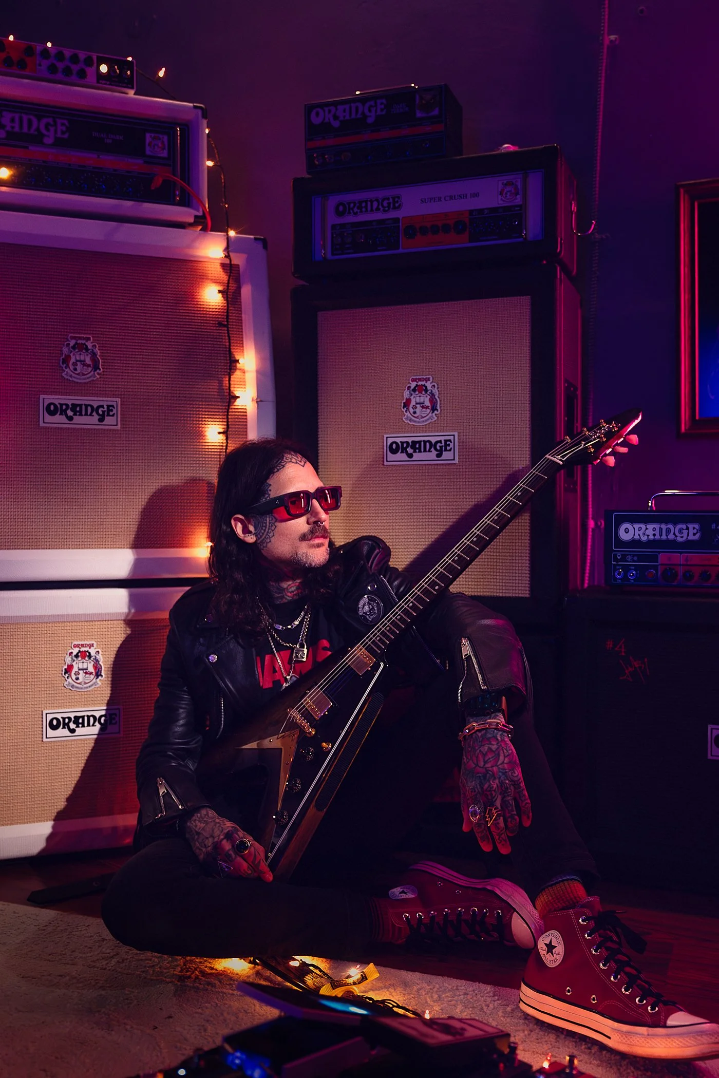 A man with tattoos and long hair, wearing sunglasses, a black leather jacket, and red Converse sneakers, sitting on the floor in front of multiple Orange amplifier stacks, holding a black electric guitar, in a dimly lit room with purple and pink ligh