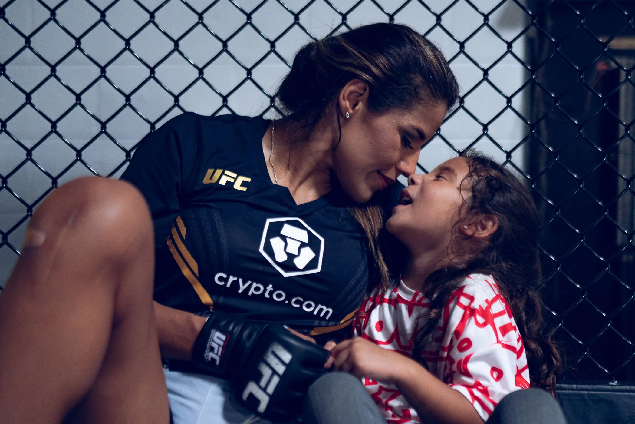 UFC Champion Julianna Pena in black fight gear and her daughter in casual clothes sharing a moment of affection inside a UFC octagon.