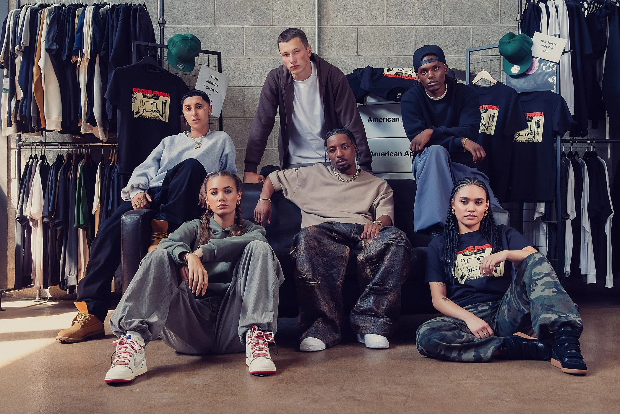 Group of six diverse young adults in casual clothing sitting and standing in a clothing store with racks of shirts and accessories behind them.