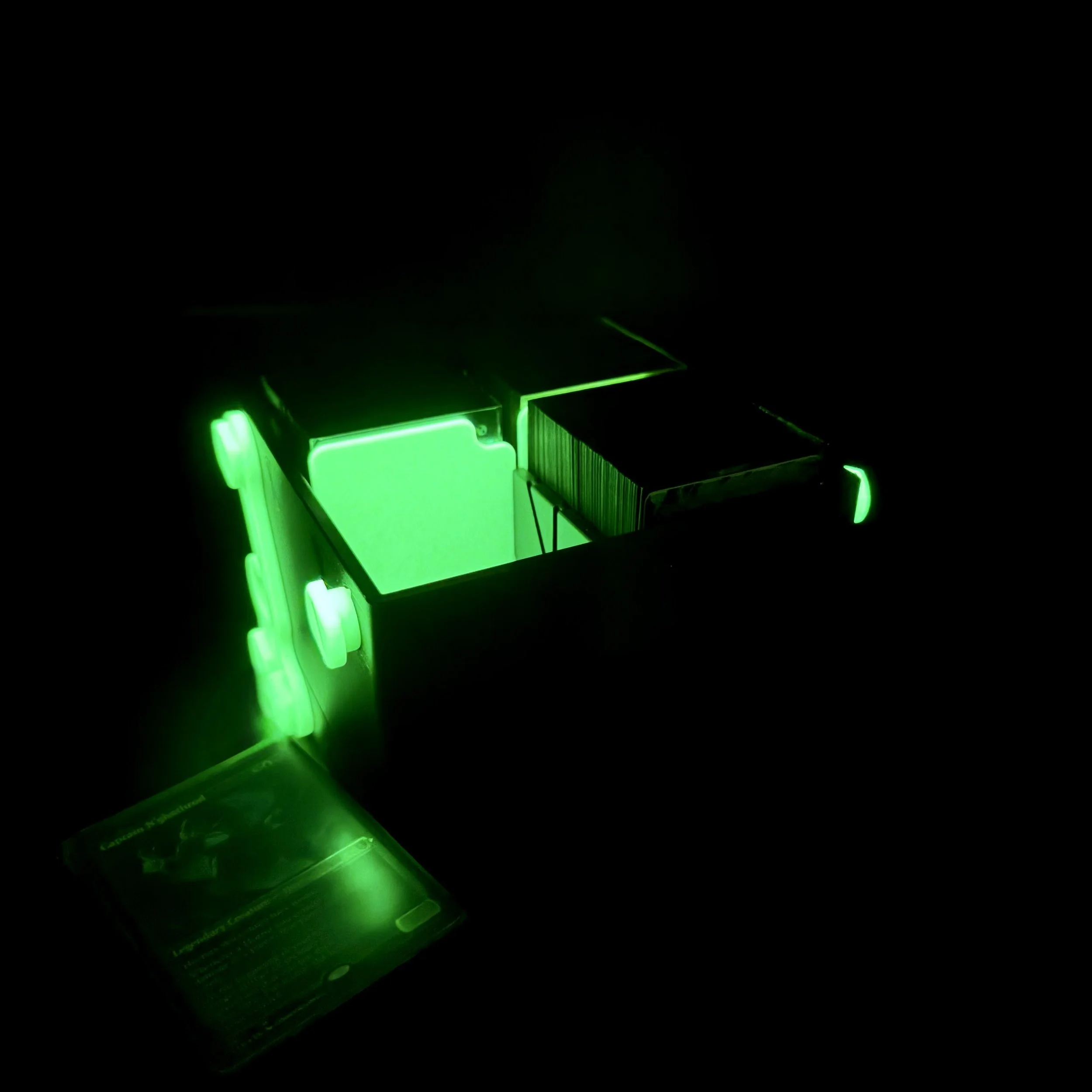 A black and glow in the dark green deckbox shown glowing with the lights off, lid stored underneath it
