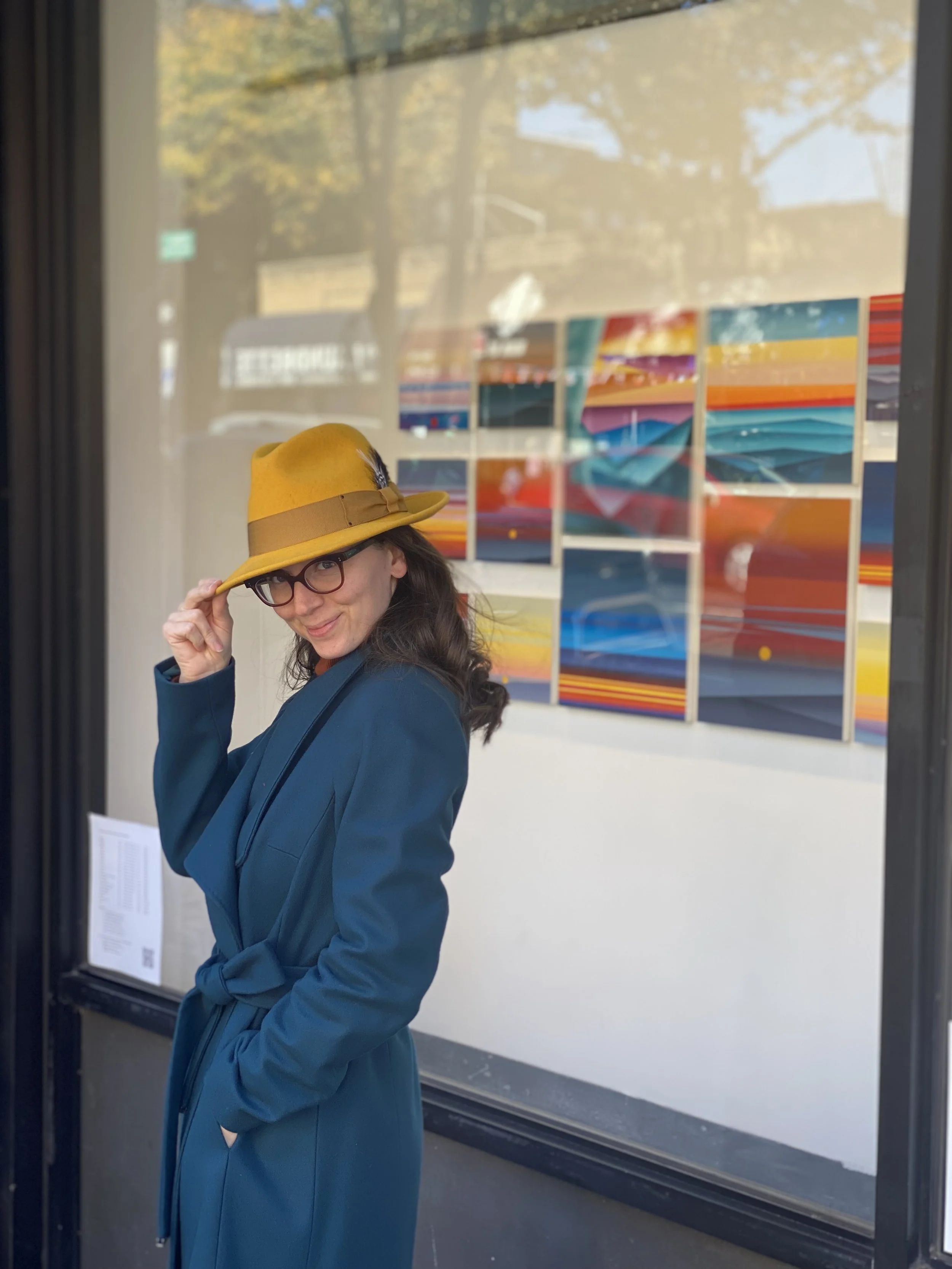 Woman in teal coat and yellow hat with feather, standing in front of an art gallery window, with colorful abstract paintings on display inside.