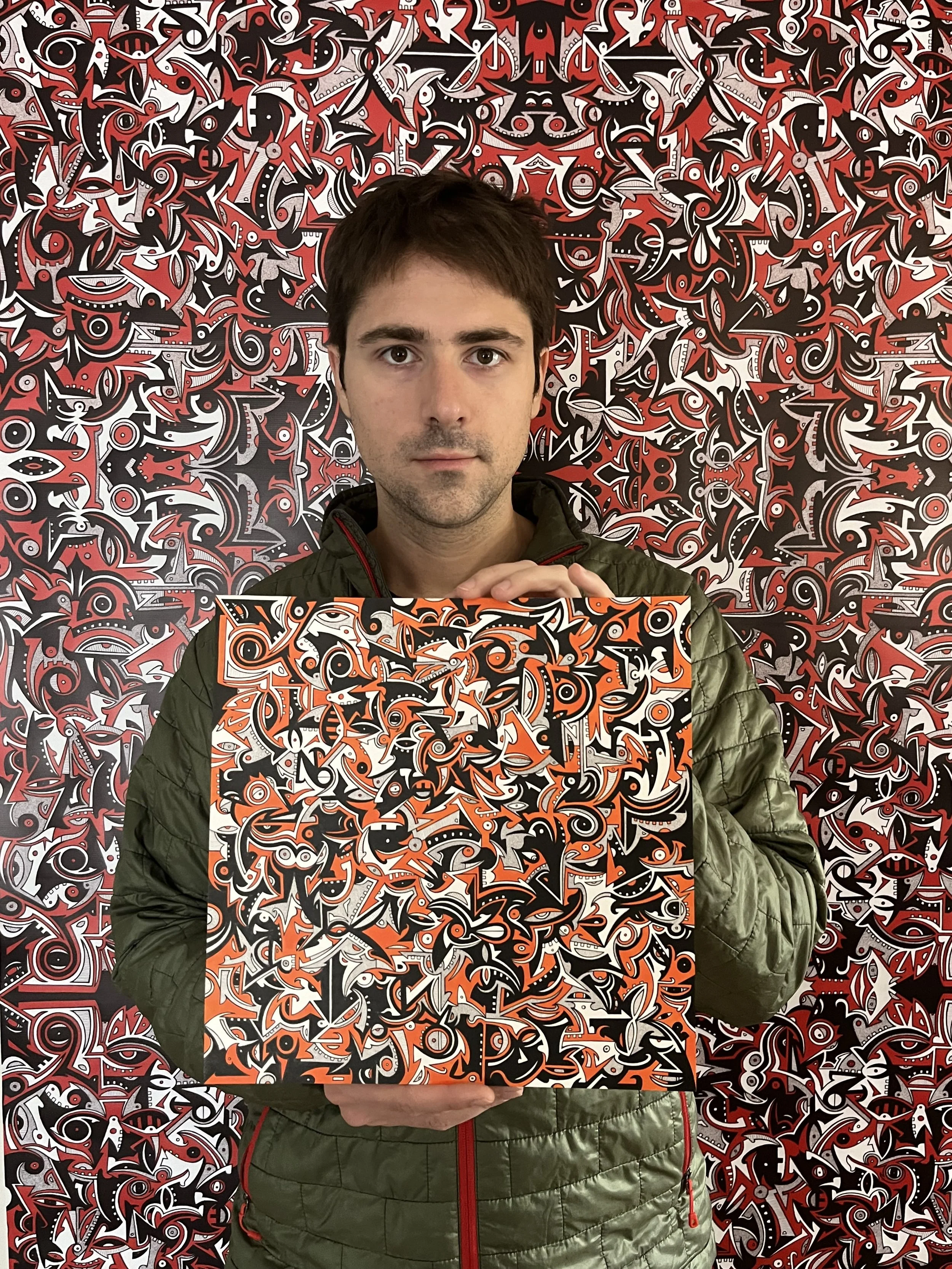 A man with short dark hair holding a square painting with a vibrant, abstract, black, white, orange, and red pattern. The background wall features a similar abstract pattern.