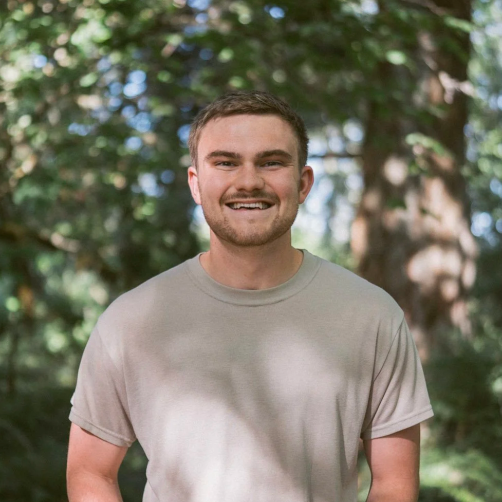 A young man with short brown hair and a beard smiling outdoors with trees and greenery in the background.