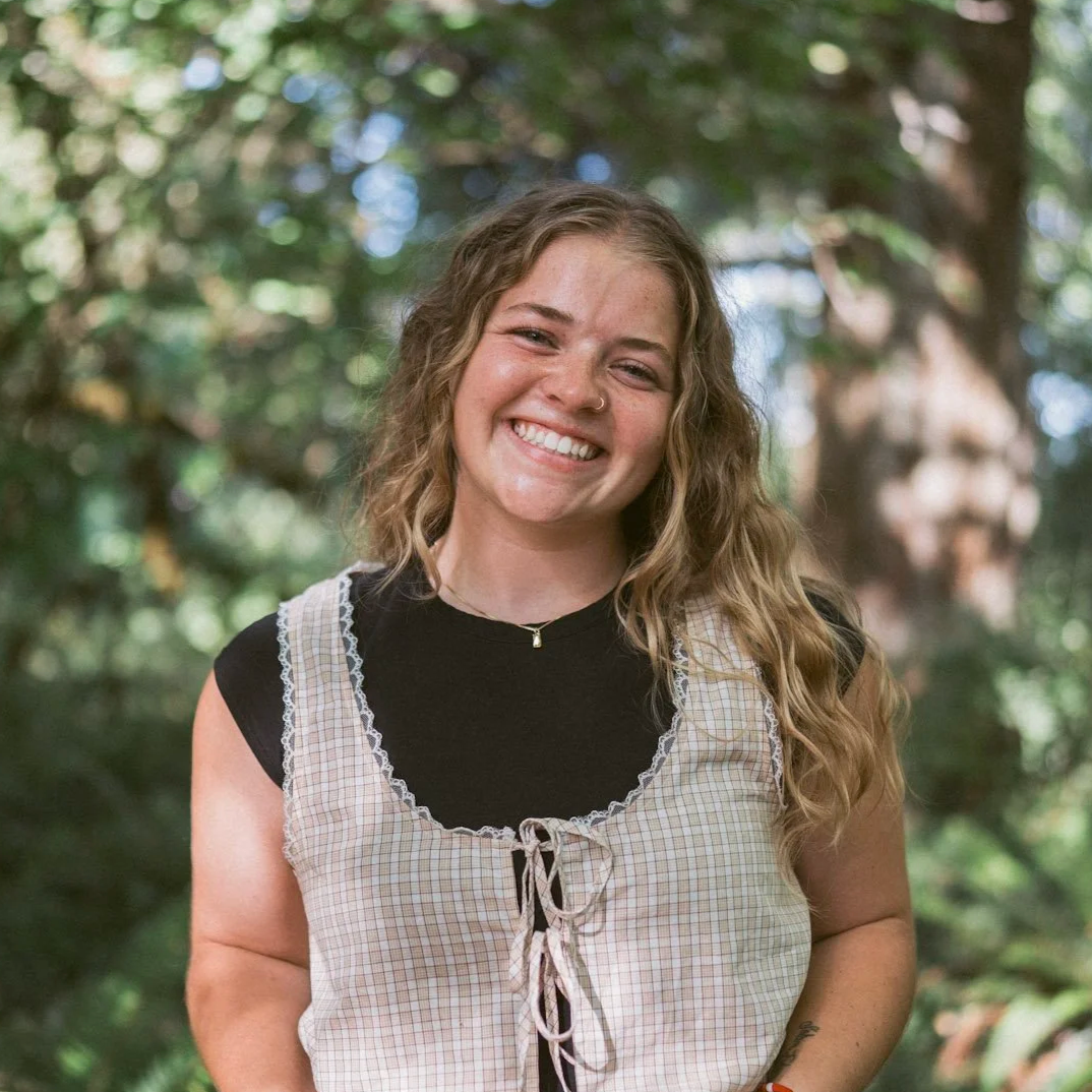 A young woman with wavy blonde hair, smiling outdoors in a wooded area, wearing a black top under a checkered sleeveless shirt with lace details.
