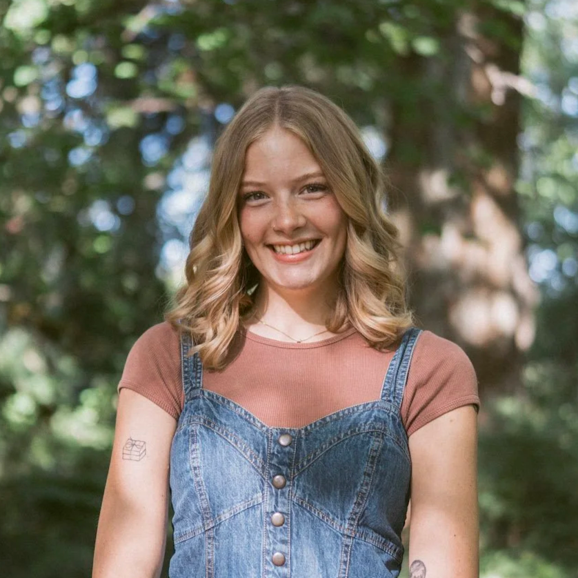 Young woman with wavy blonde hair smiling outdoors, wearing a brown t-shirt under a denim overall. Background features green trees and sunlight.