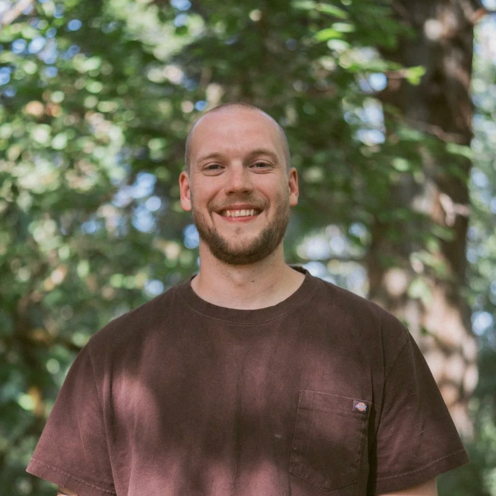 A smiling man with a beard and short hair standing outdoors in front of a tree with green leaves.