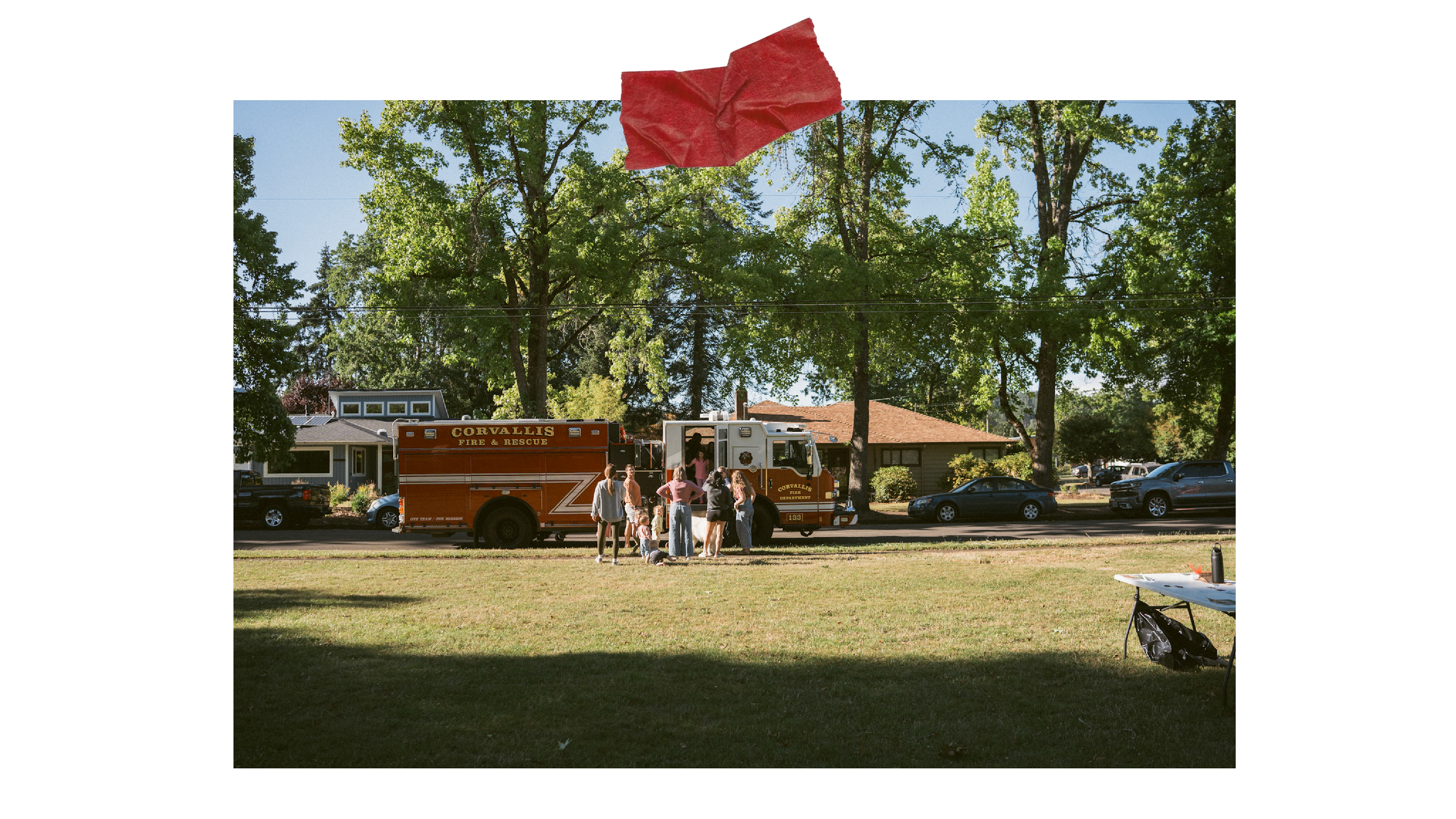 Firefighters and residents gather near a fire truck in a neighborhood park, with a large tree and residential houses in the background on a sunny day.