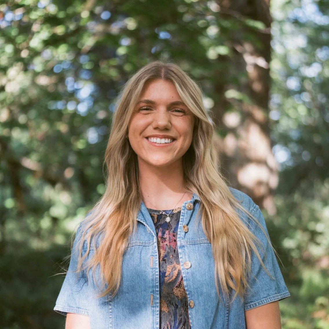 Young woman with long blonde hair smiling outdoors, wearing a denim jacket over a patterned top, with trees and foliage in the background.