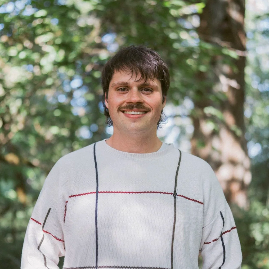 Man smiling outdoors, wearing a white sweater with colorful patterns, trees in the background.