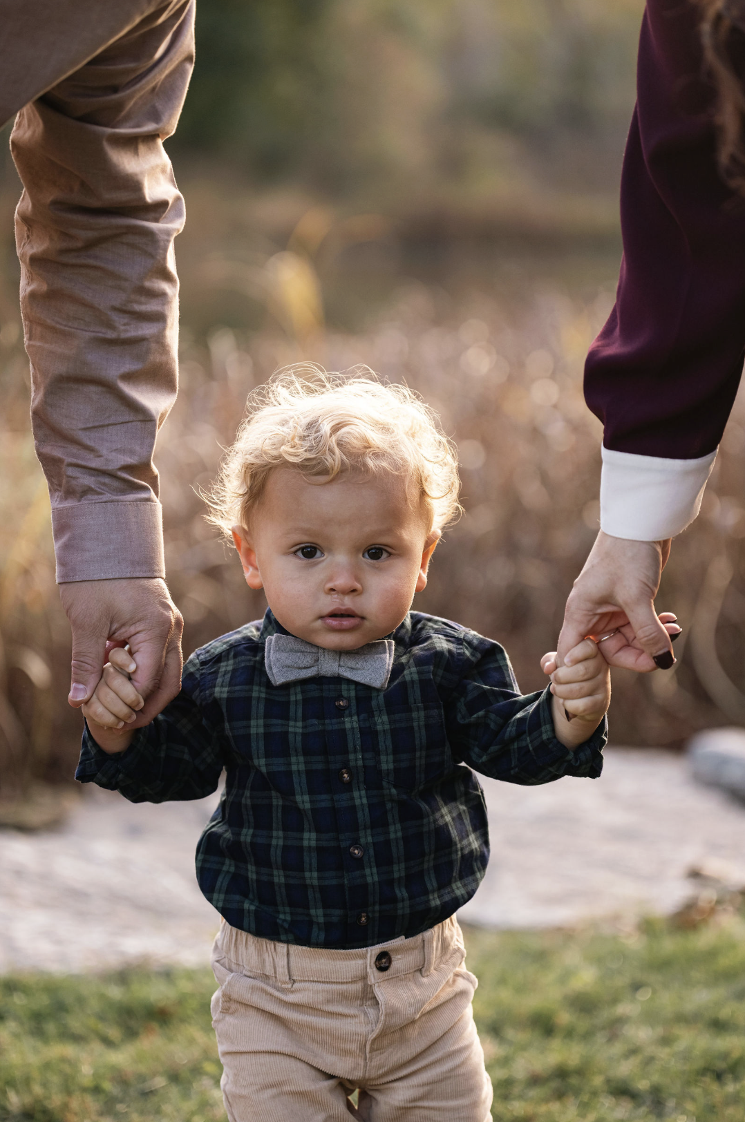 A young boy with curly blonde hair wearing a plaid shirt, beige pants, and a bow tie is holding hands with two adults, walking outdoors in a natural setting with soft sunlight and blurred foliage in the background.