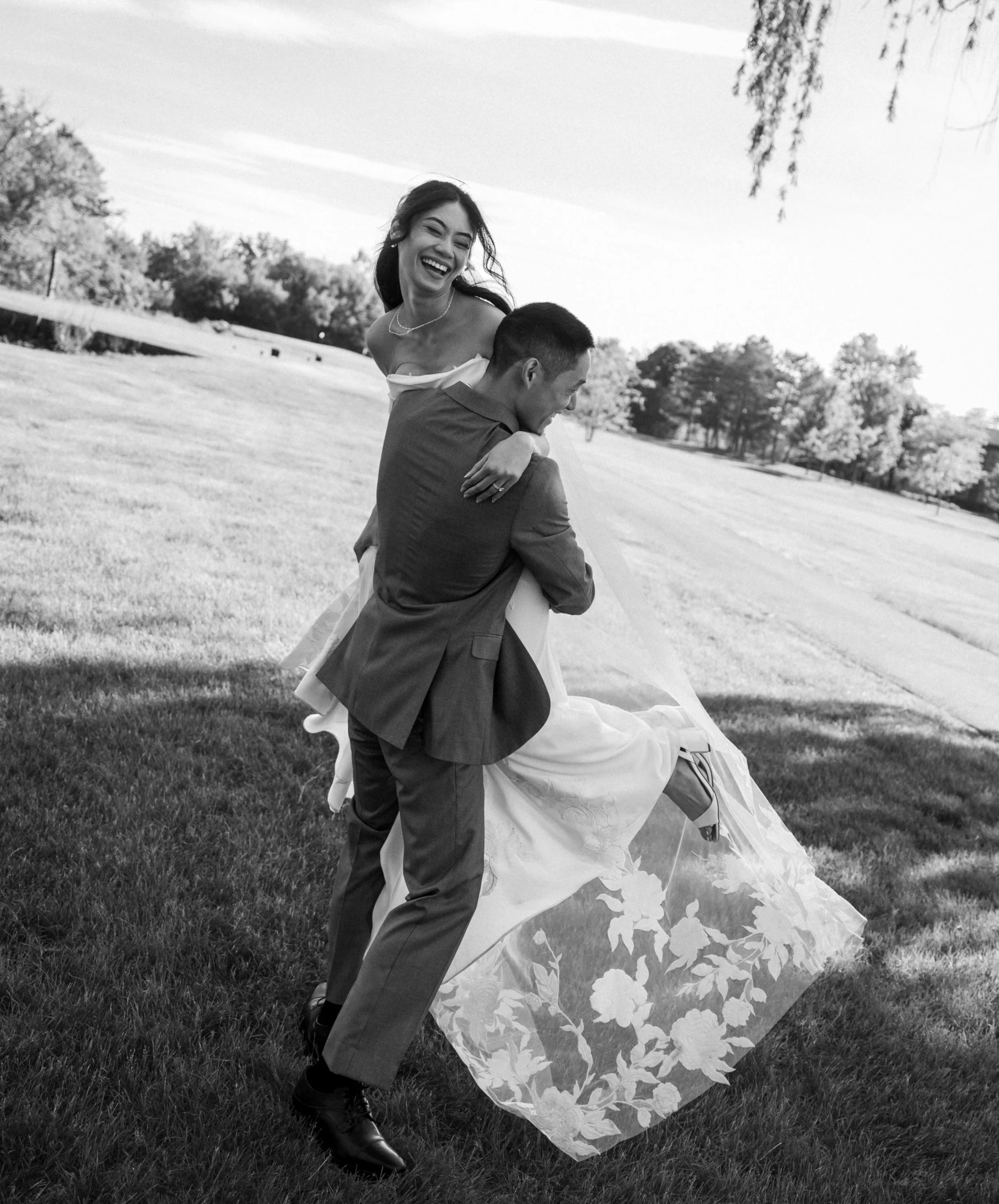 A woman in a wedding dress being carried by a man outdoors on a grassy field, laughing and smiling. Black and white photograph with trees in the background.