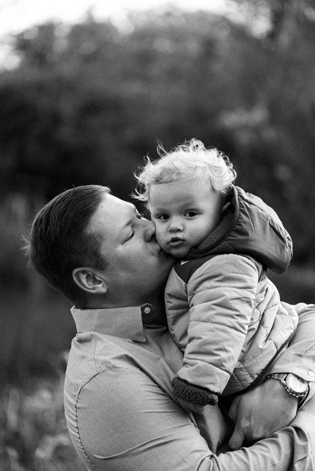 A man kisses a young child on the cheek while holding him outdoors, with a blurred natural background.