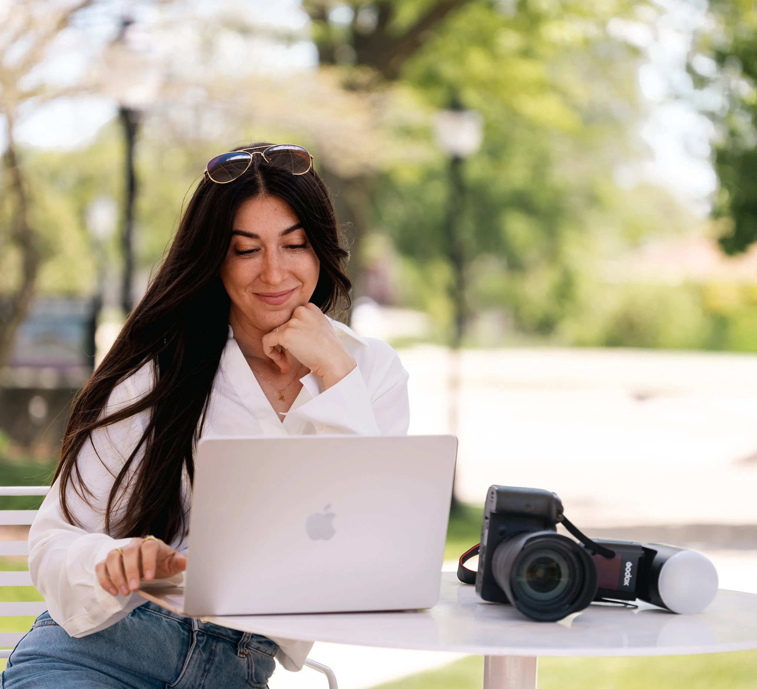 Young woman with sunglasses on her head sitting at a table outdoors using a laptop, with camera equipment nearby.