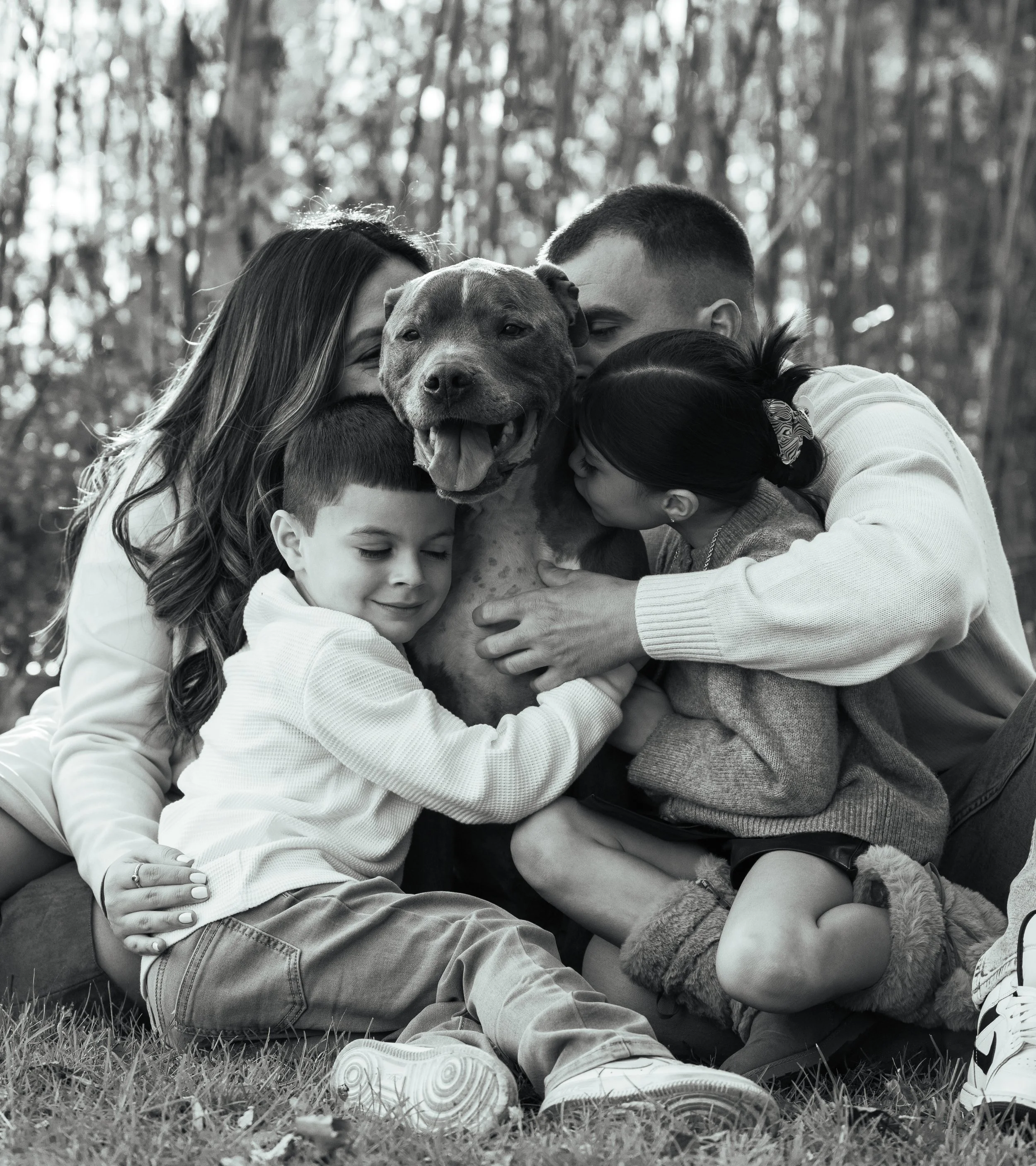 Black and white photo of three children hugging a large dog outdoors. The children are smiling and appear happy, with sunlight filtering through trees in the background.
