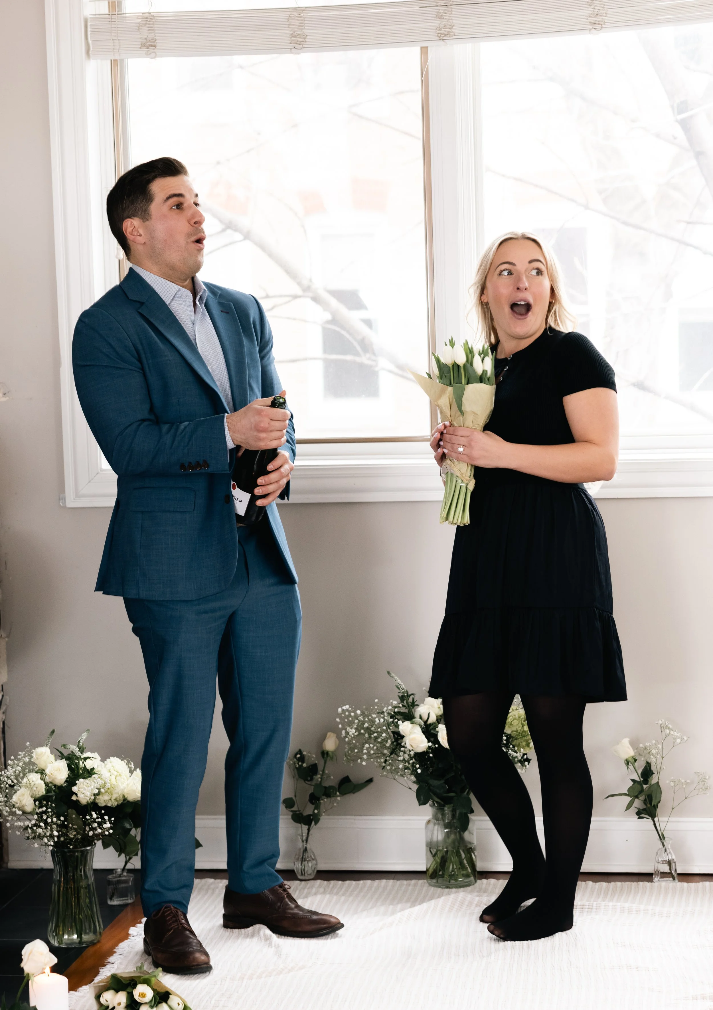 A man in a blue suit presents a bottle of champagne to a woman holding a bouquet of white tulips. They are standing in front of a large window with white curtains, surrounded by white flowers and candles, in a room decorated for a celebration or spec