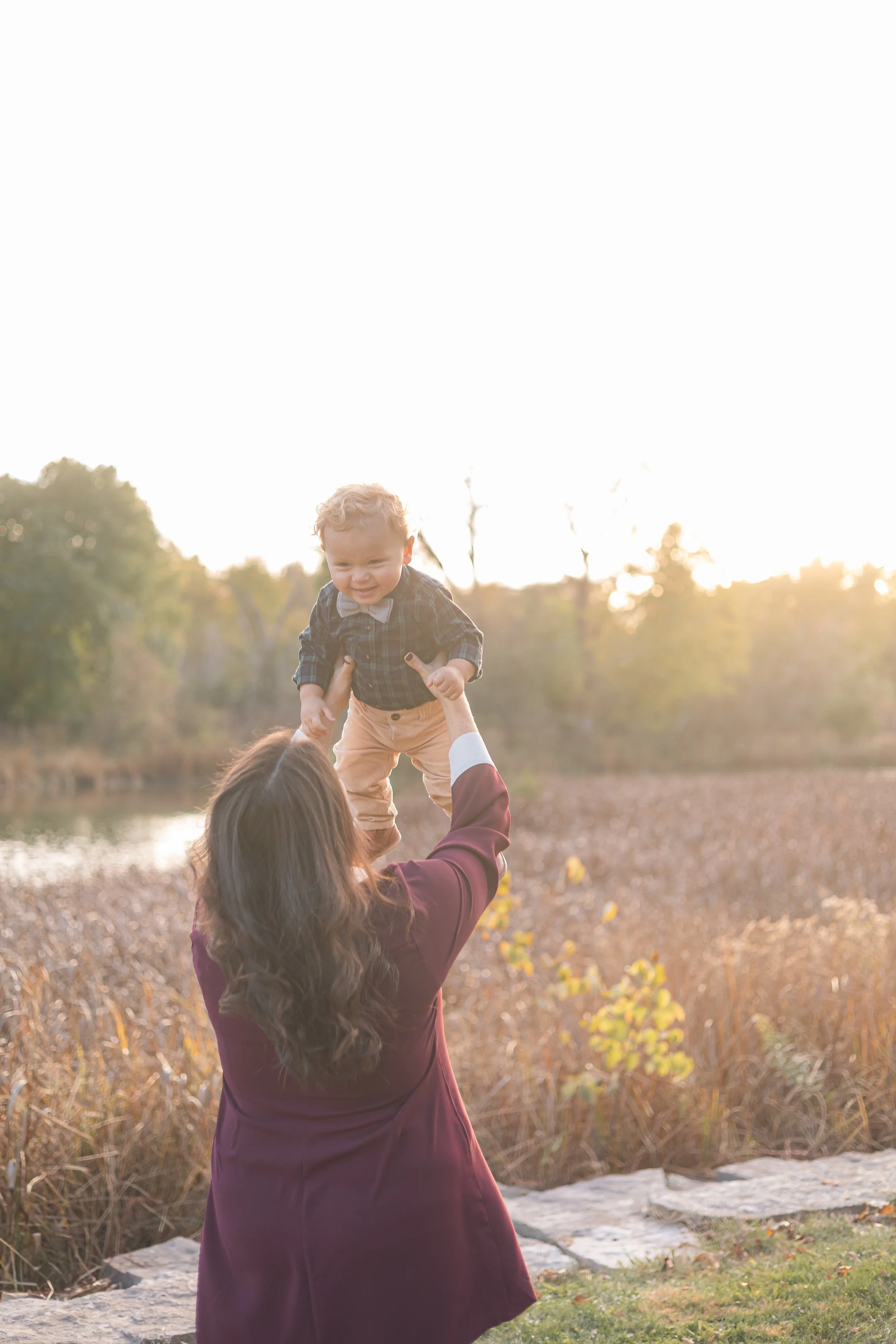 A woman with long dark hair, wearing a burgundy coat, is lifting a young boy in a plaid shirt and beige pants into the air outdoors during sunset. They are near a body of water and surrounded by trees and autumn foliage.