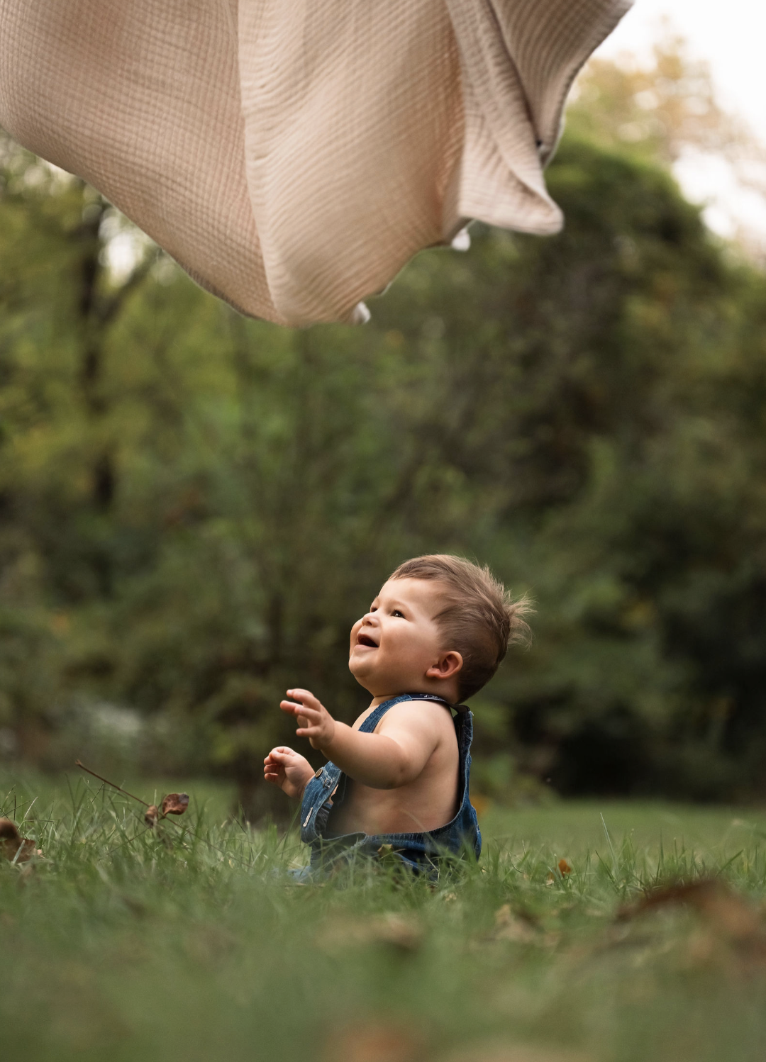 A young child sitting on grass looking up and smiling at a large beige fabric structure hanging overhead outdoors with trees in the background.