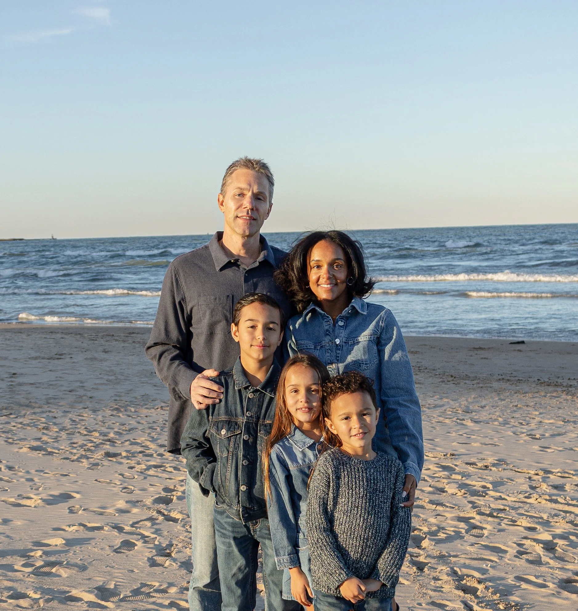 Family of seven standing on a sandy beach with ocean waves in the background during sunset.