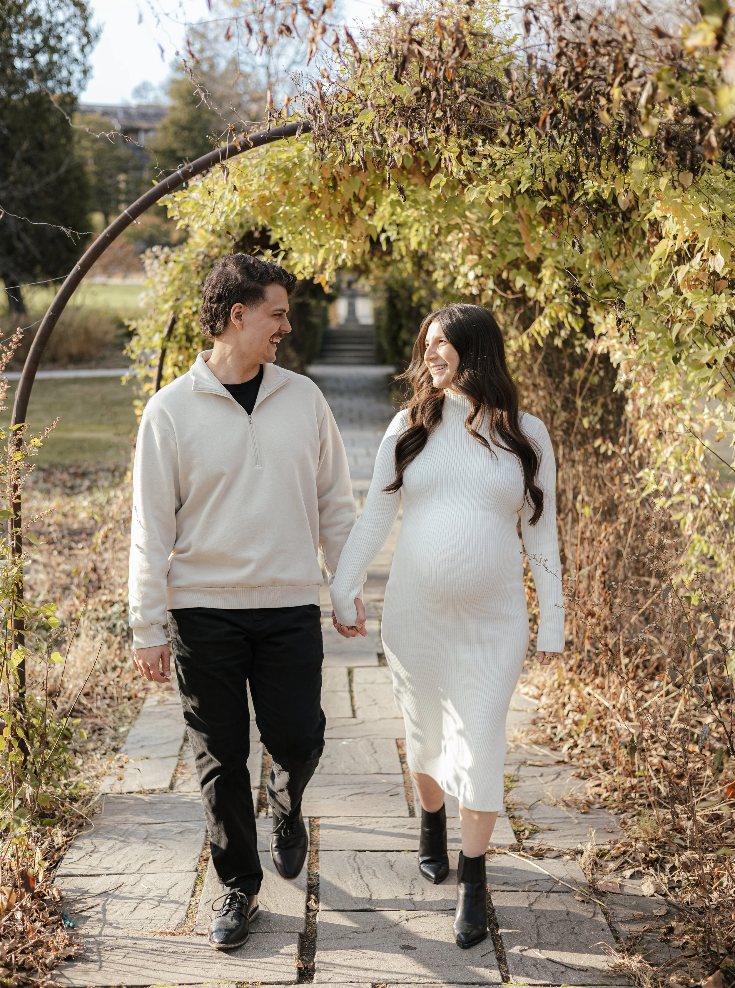 A smiling young man and woman walking hand in hand through a park archway covered with autumn leaves. The man wears a beige zip-up sweater and black pants; the woman wears a white ribbed dress and black ankle boots. They look at each other happily.