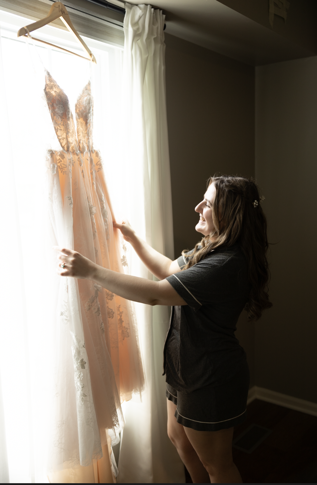 A woman smiling as she looks at a peach-colored wedding dress with lace details hanging in front of a window.