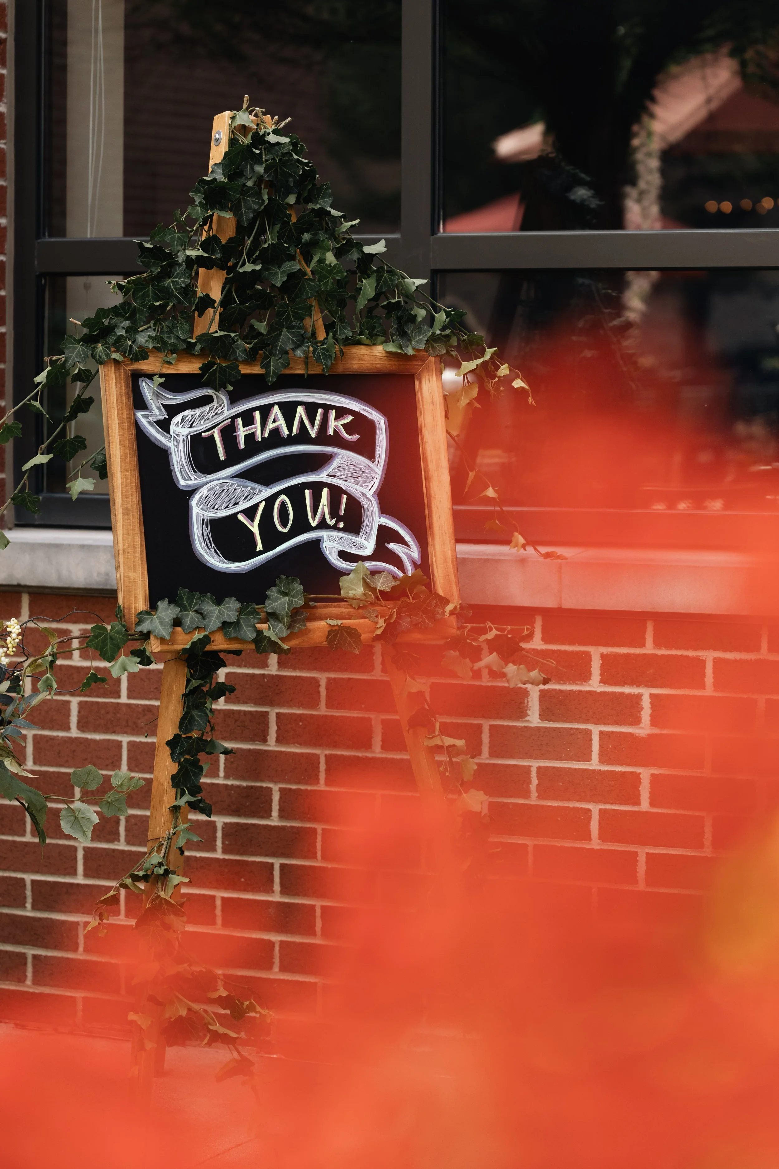A chalkboard sign decorated with green ivy leaves, displaying a neon-style message that says "Thank You!" The sign is positioned outside near a brick wall and a window, with a soft light illuminating the scene.