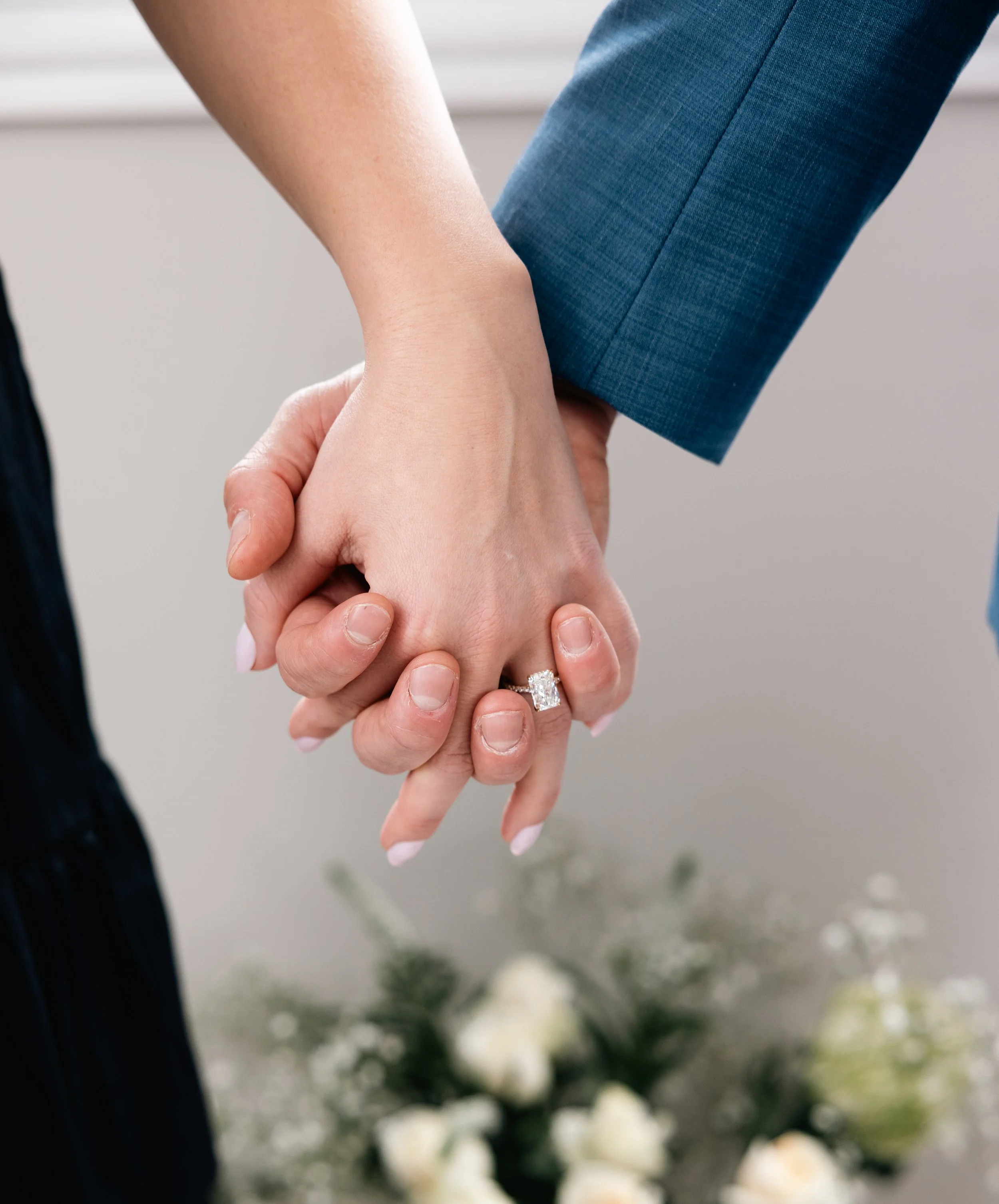 A person in a suit holding hands with a woman, showcasing an engagement ring, with blurred flowers in the background.