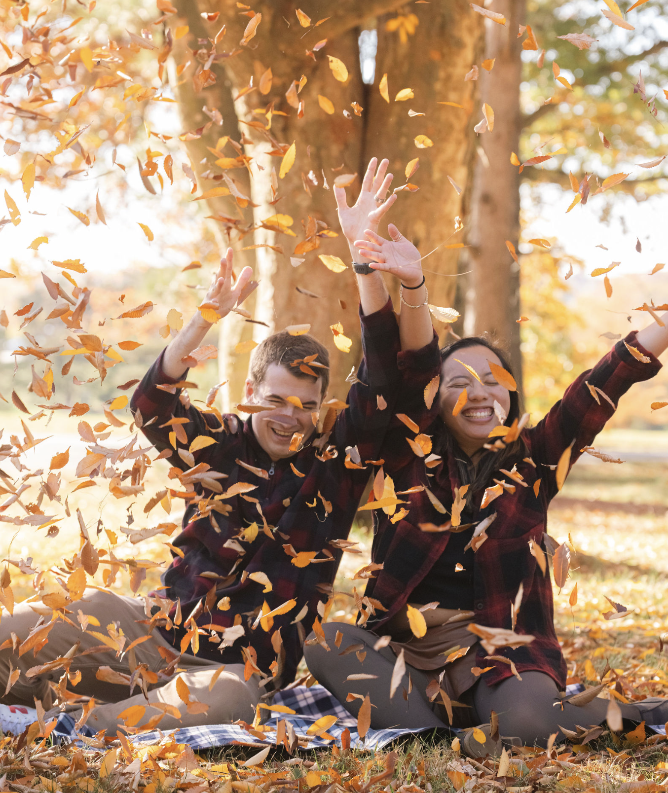 Two people sitting on a blanket outdoors during autumn, throwing orange and yellow leaves in the air, smiling and enjoying the fall weather with trees in the background.