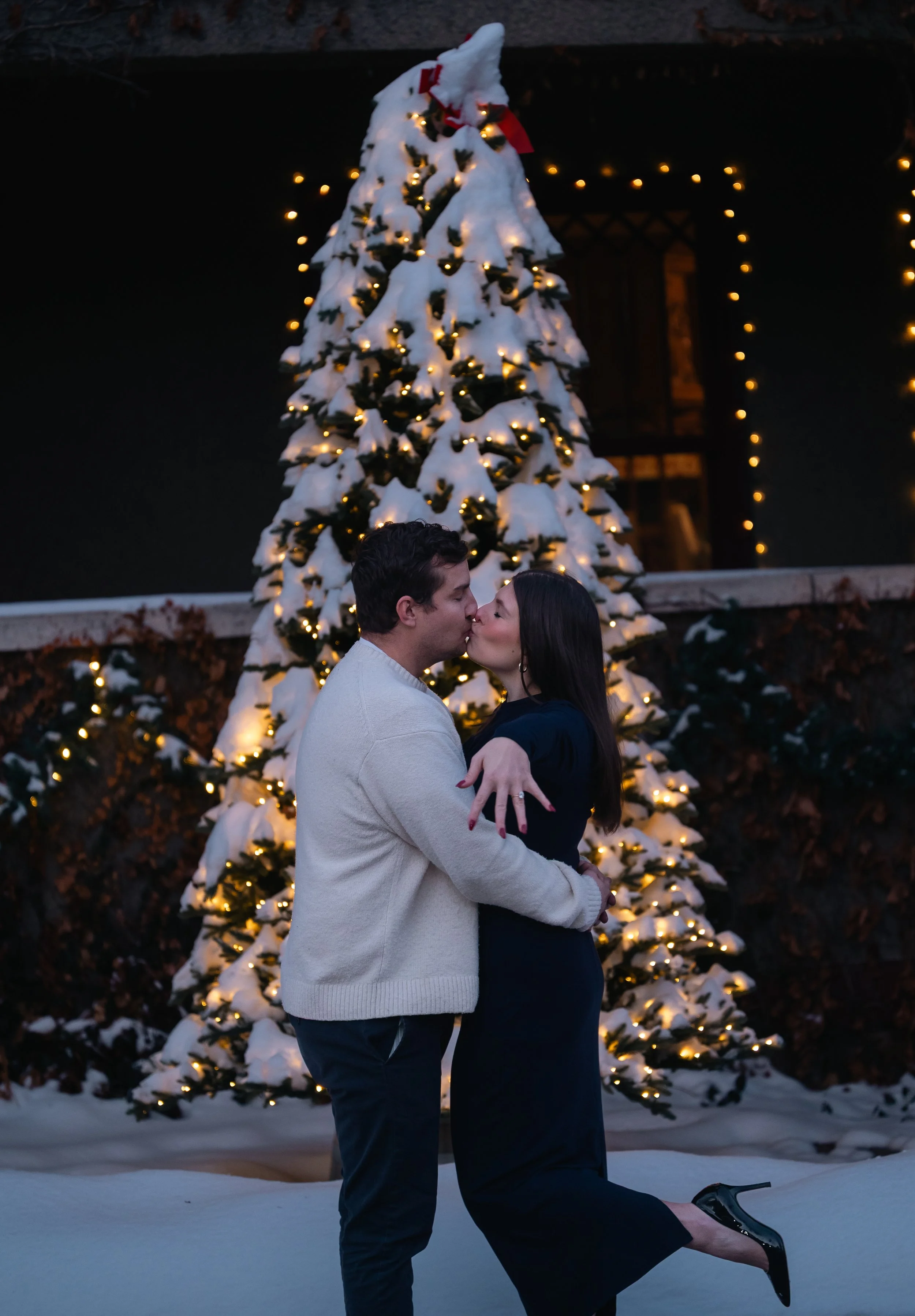 A couple kissing outdoors in front of a snow-covered Christmas tree with lights, during nighttime.