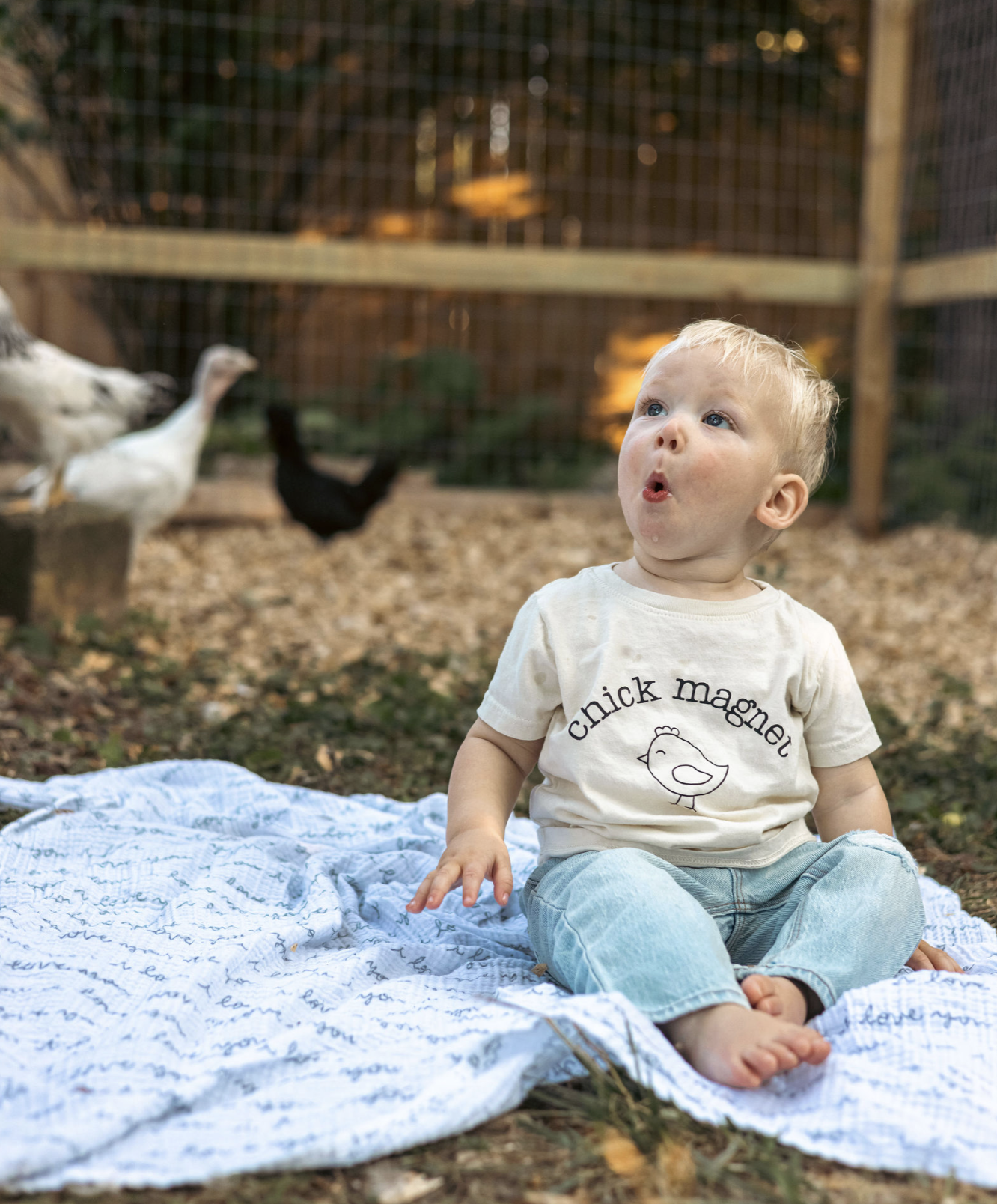 A young child sitting on a blanket outdoors near a chicken coop, with chickens in the background. The child is wearing a cream-colored shirt with a chicken illustration and the words 'chick magnet' on it, and light blue pants. The child has blonde ha