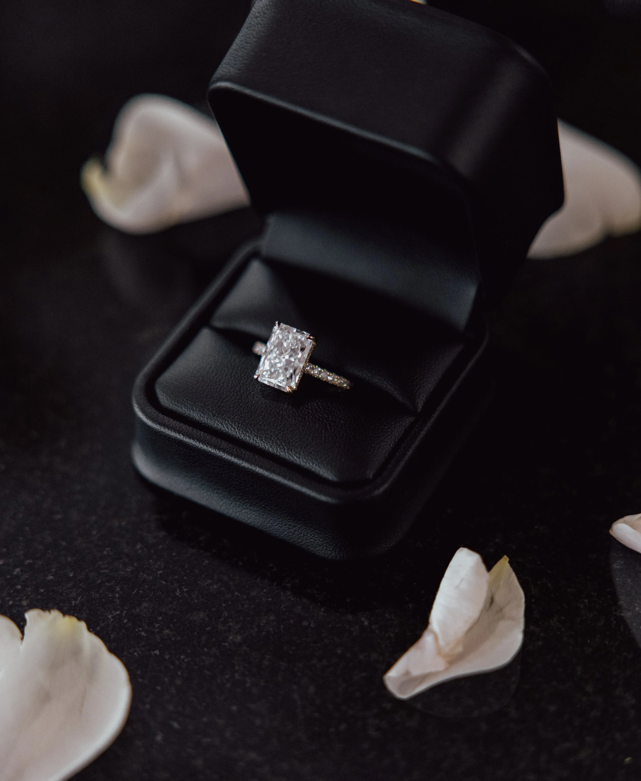 A black jewelry box with a large rectangular diamond ring inside, surrounded by scattered white seashells on a dark surface.