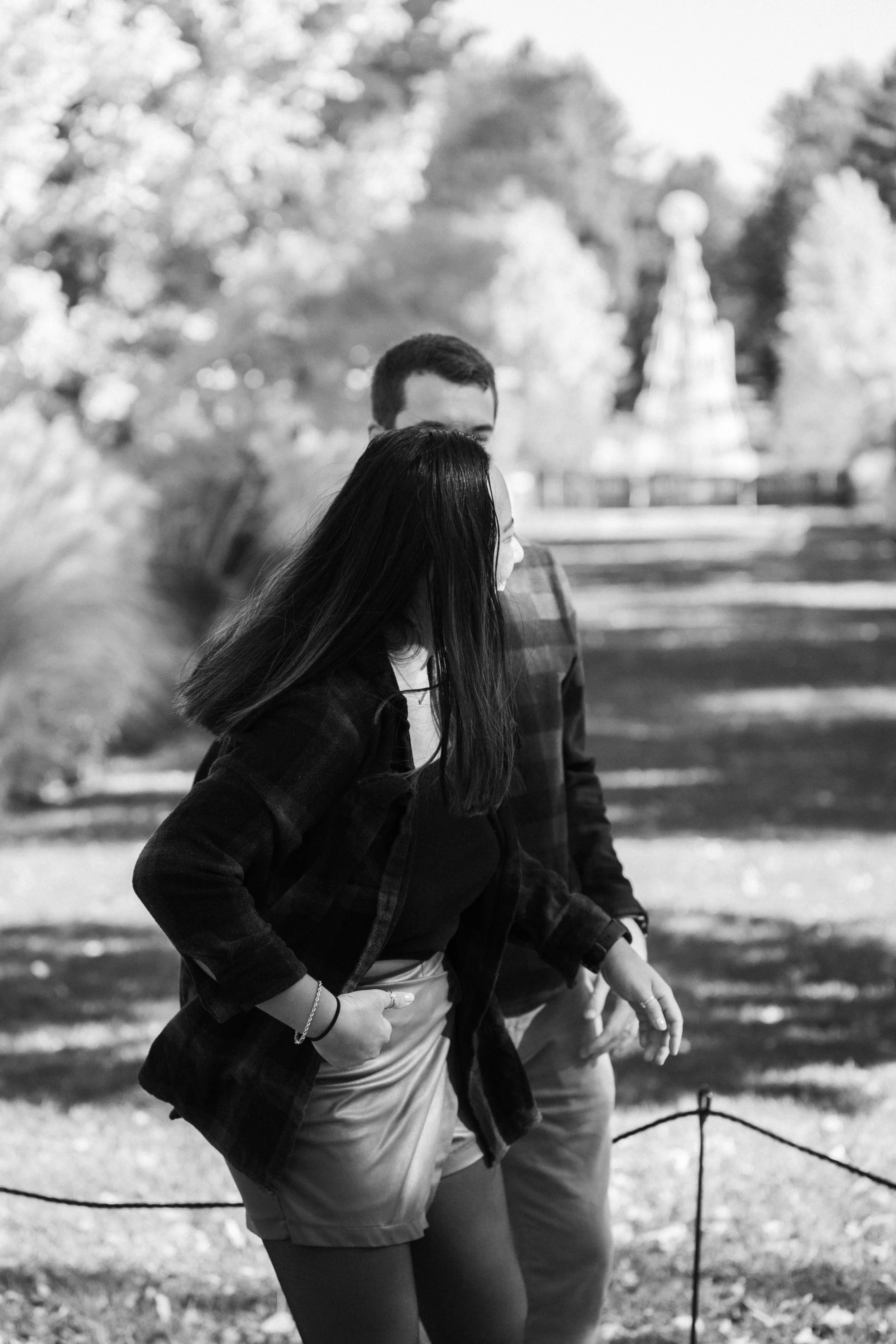 A black and white photo of a woman walking outdoors, with trees and a structure in the background.