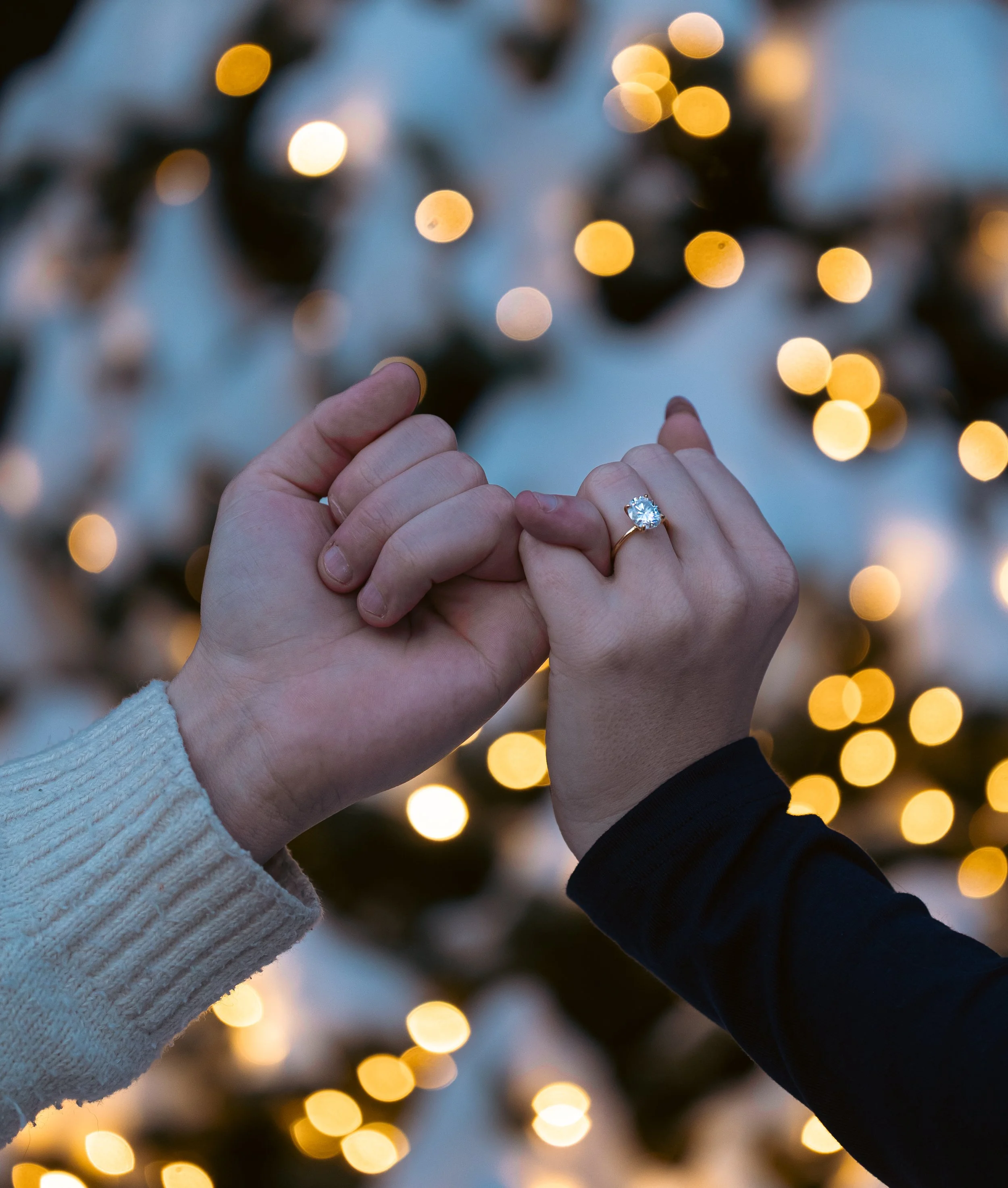 Two hands, one with a large diamond ring, are intertwined in front of a blurred background of golden holiday lights.