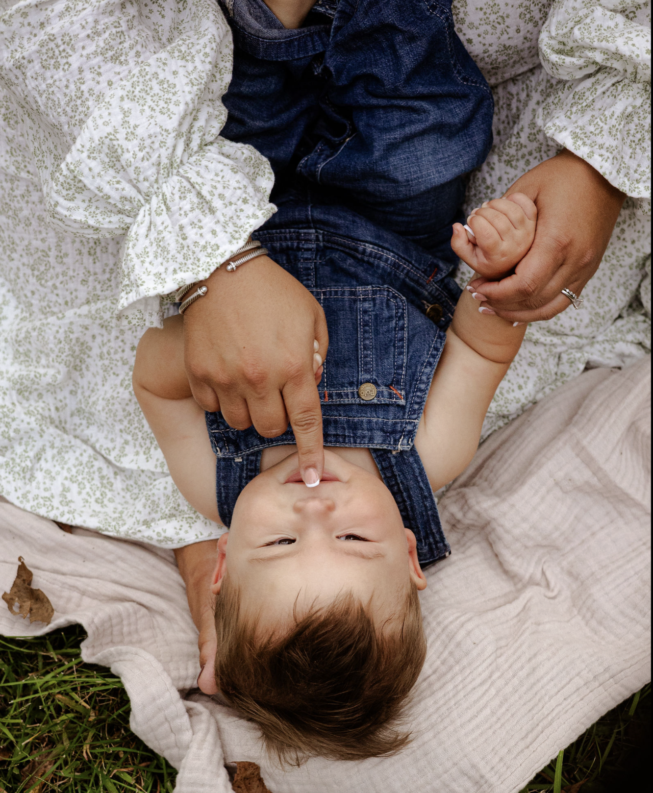 A young child with brown hair laying on a bed of leaves and grass on a beige blanket. The child is wearing a denim vest and blue jeans, with one finger pressed to their nose. An adult's hands are holding and gently pressing the child's finger.