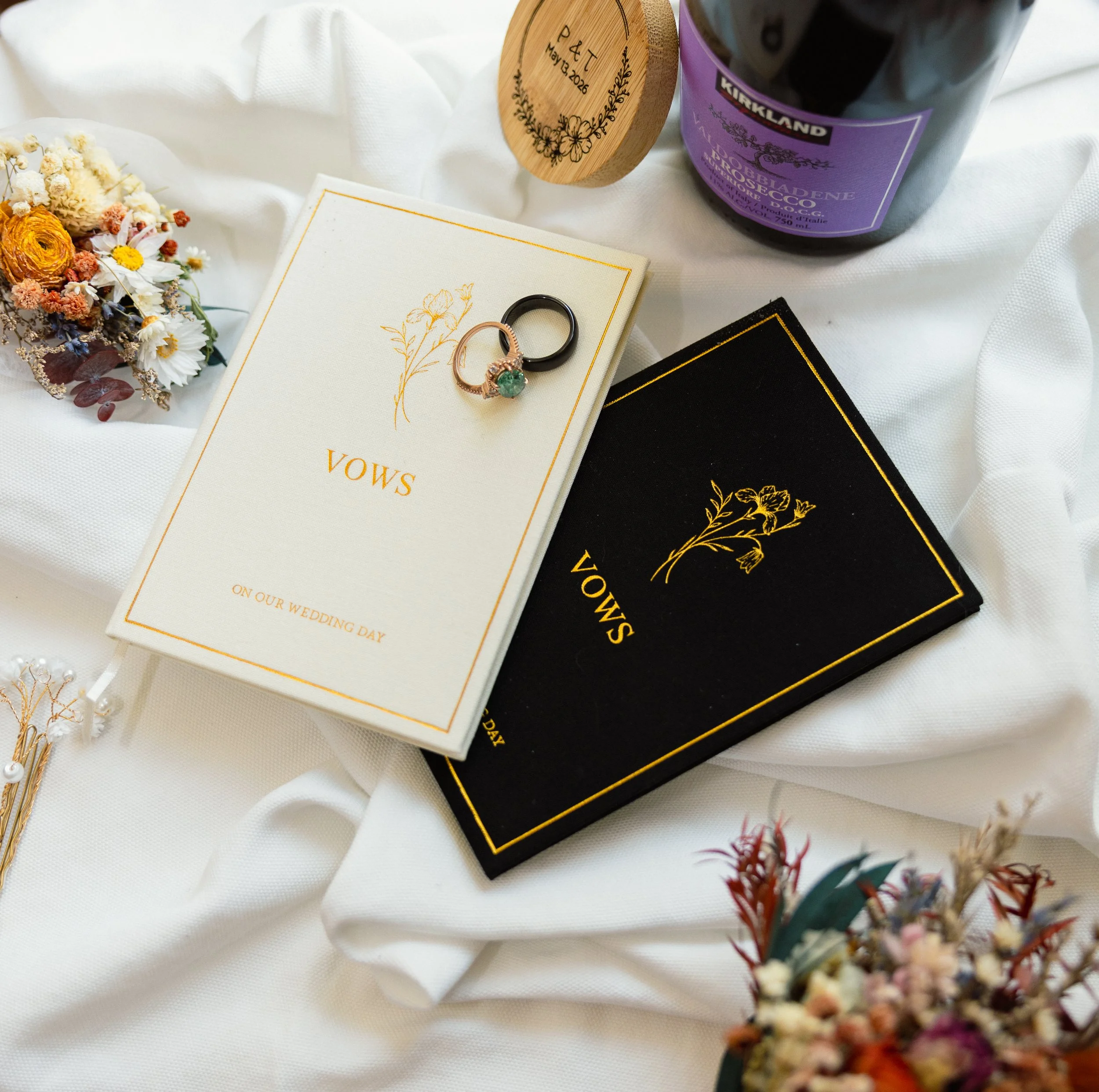 A wedding ring with a green gemstone on a white card, alongside a dried flower bouquet, a purple bottle of Kirkland red wine, a black velvet box, and a floral arrangement on a white fabric background.