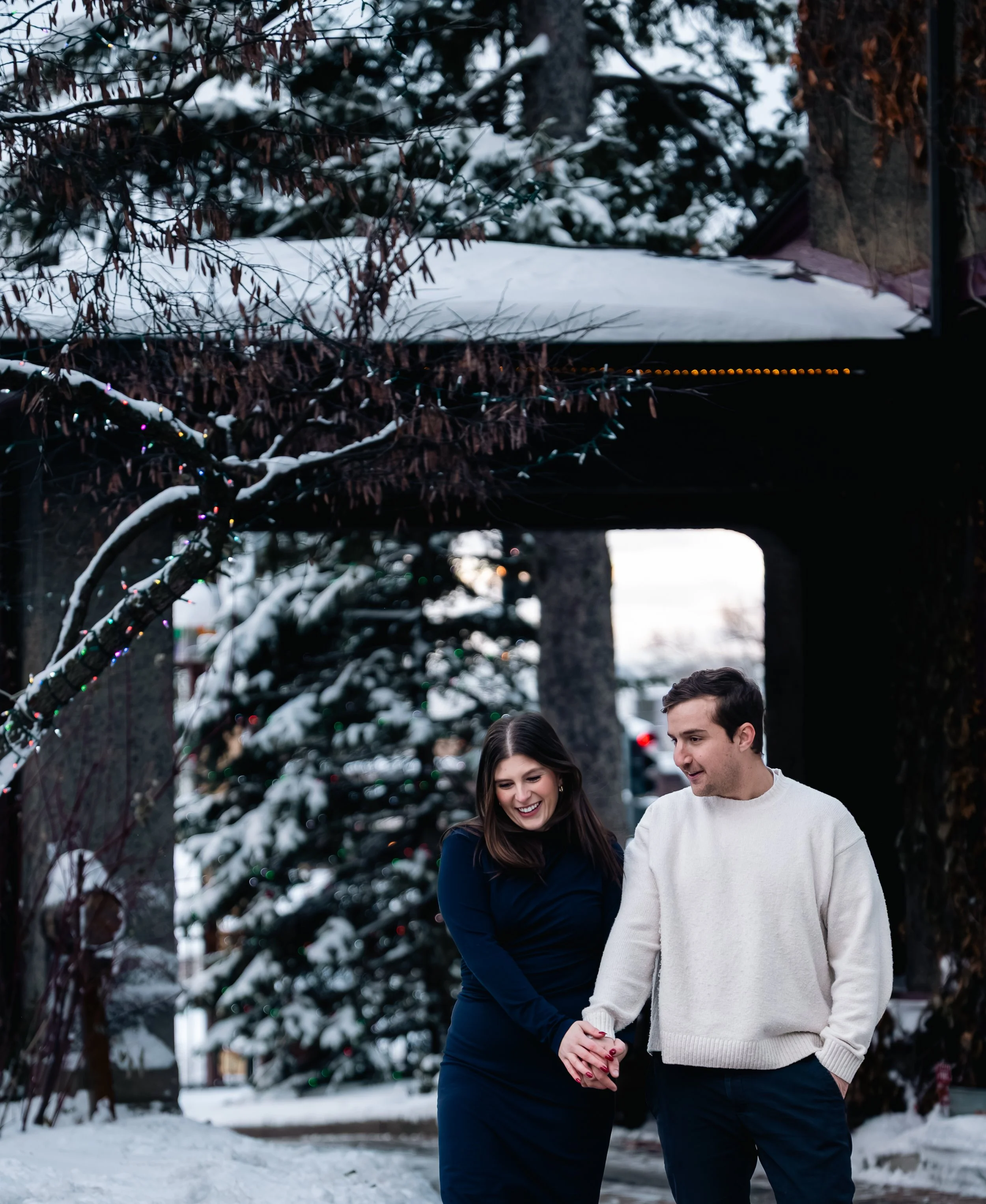 A smiling couple, a woman and a man, holding hands and walking outdoors in a snowy setting with a decorated Christmas tree and snow-covered ground.