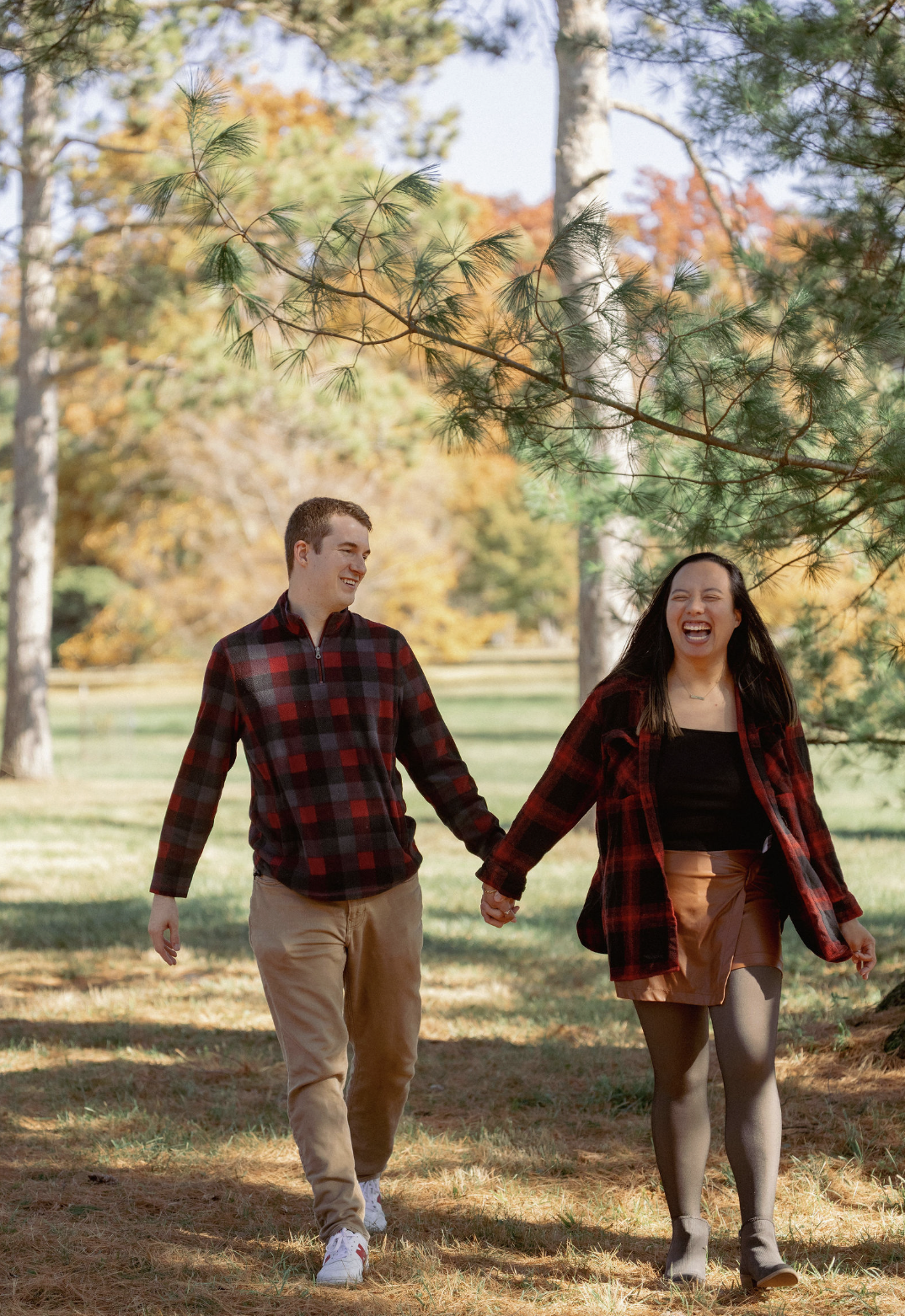 Two young adults, a man and a woman, holding hands and walking through a park with fall foliage, both wearing red plaid shirts, smiling and enjoying the autumn day.