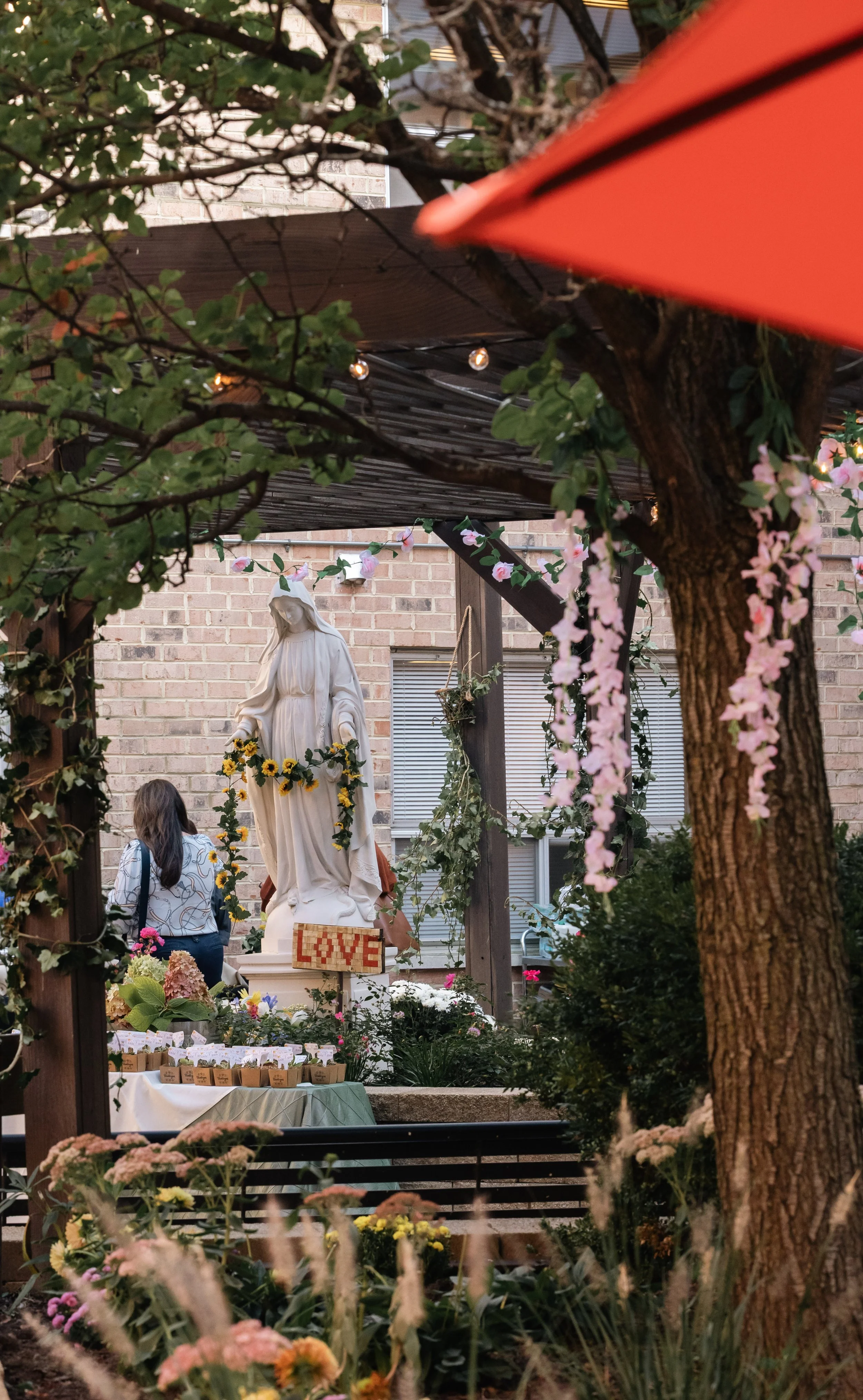 A garden scene with a statue of Jesus Christ adorned with a sunflower crown, surrounded by pink and white flowers, plants, and a table with small potted plants, in front of a brick building.