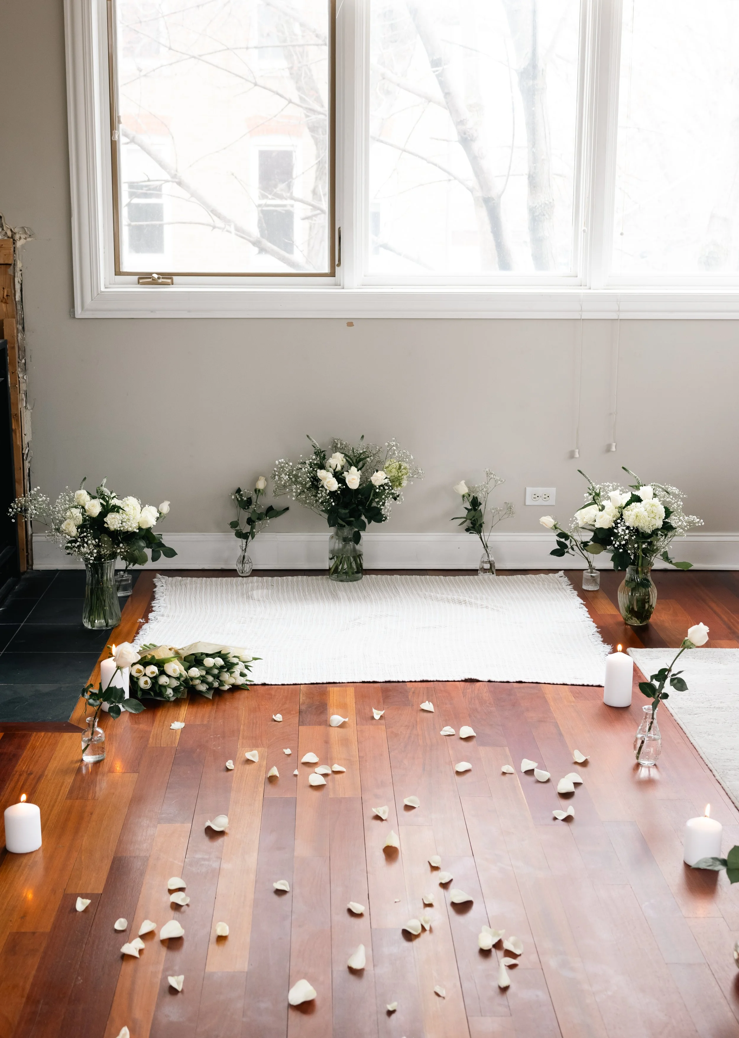 Interior of a room decorated with white flowers in vases, white candles, and rose petals scattered on a wooden floor, with a large window in the background.