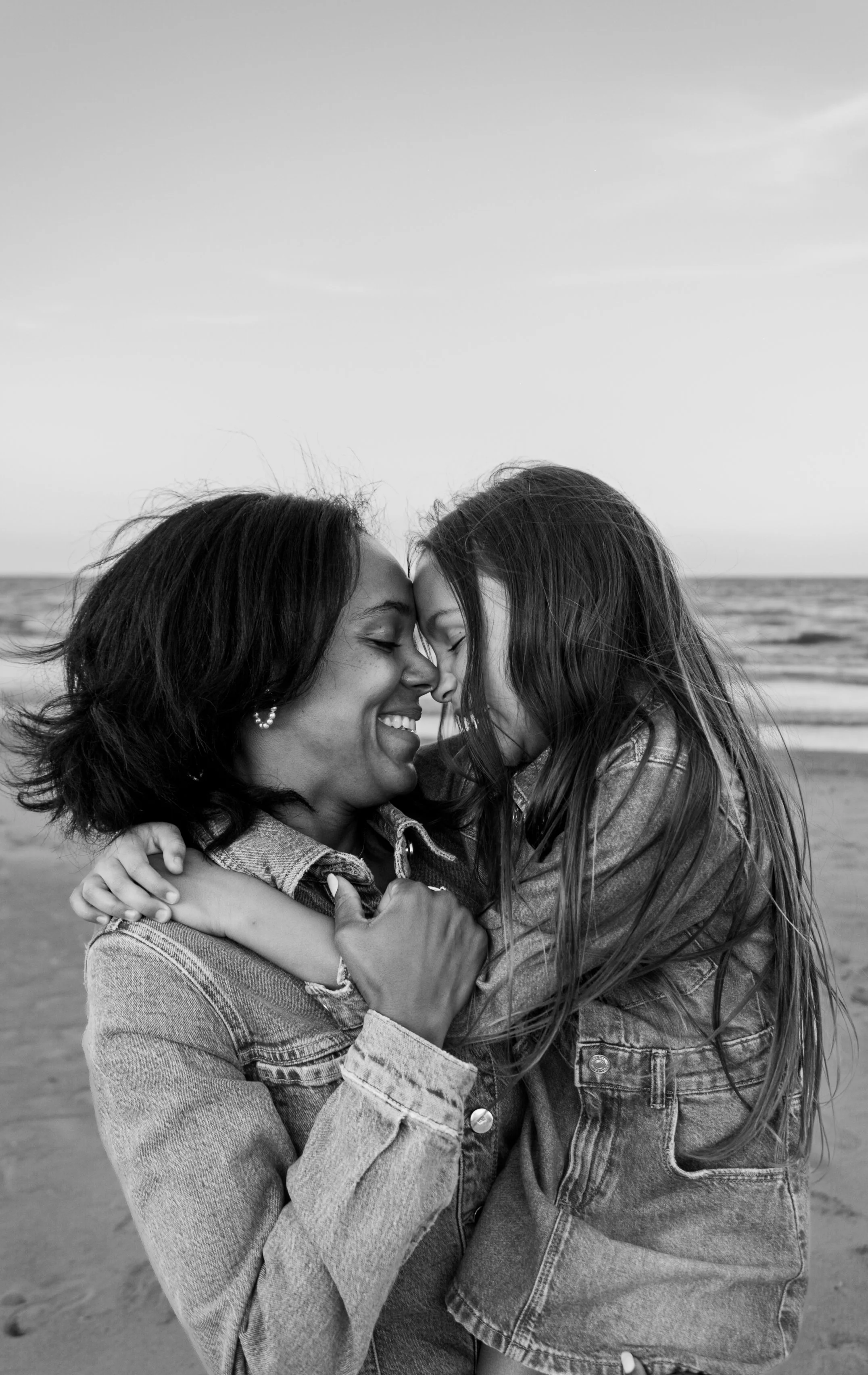 A black and white photo of two women hugging and touching foreheads, smiling with eyes closed on a beach.