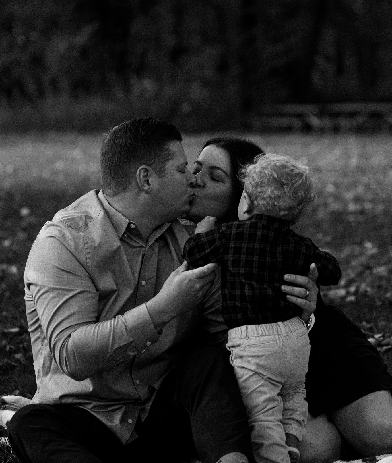 A black and white photo of a man and a woman kissing, with a young child in their arms, outdoors in a park.