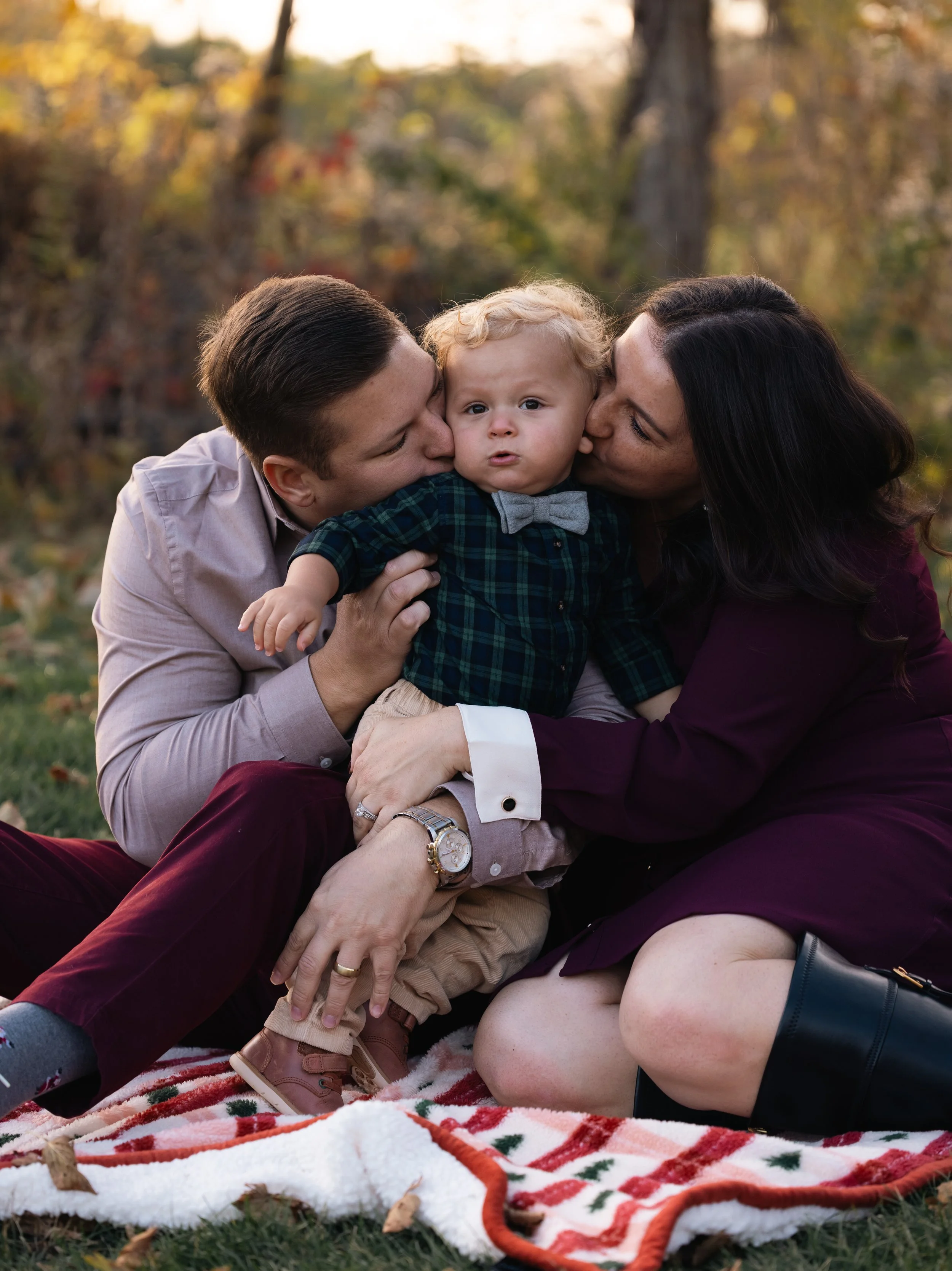 A family of three, a man, a woman, and a young boy, sitting on a blanket outdoors during autumn. The man kisses the boy's cheek, and the woman kisses the boy's other cheek. The boy looks at the camera with a slightly surprised expression. Trees with 