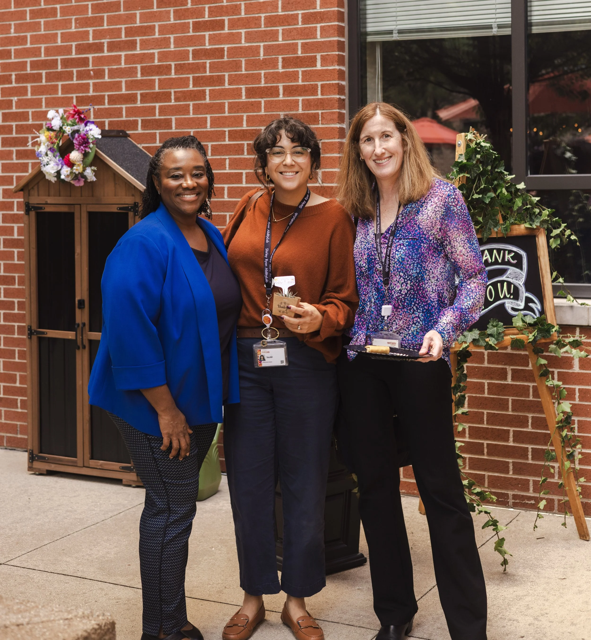 Three women standing in front of a brick wall, smiling at the camera, with a flower-adorned doghouse to their left and a chalkboard sign to their right outside a building.