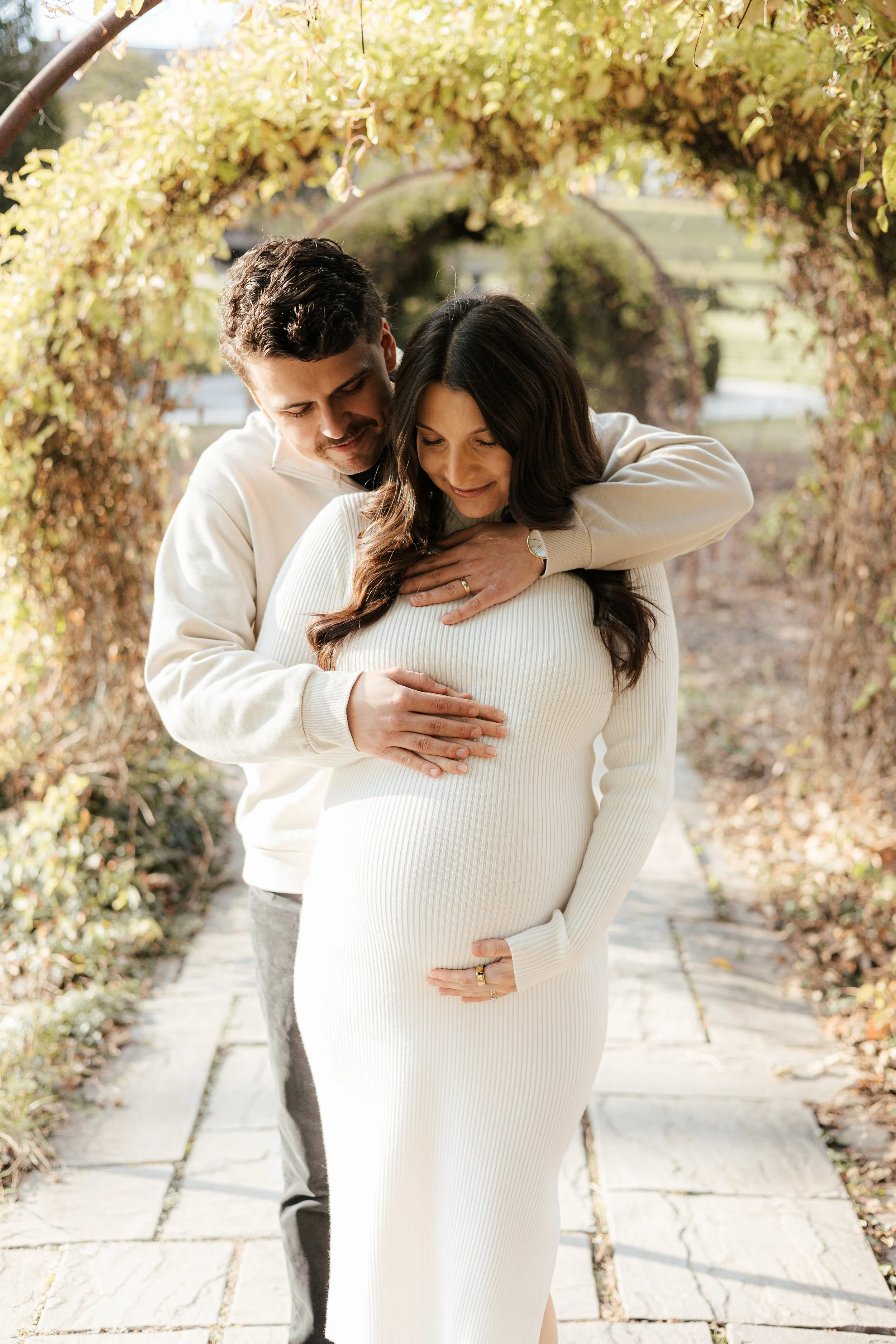 A man and woman embrace outdoors on a paved pathway surrounded by greenery and arching foliage, with the woman visibly pregnant, both smiling softly.